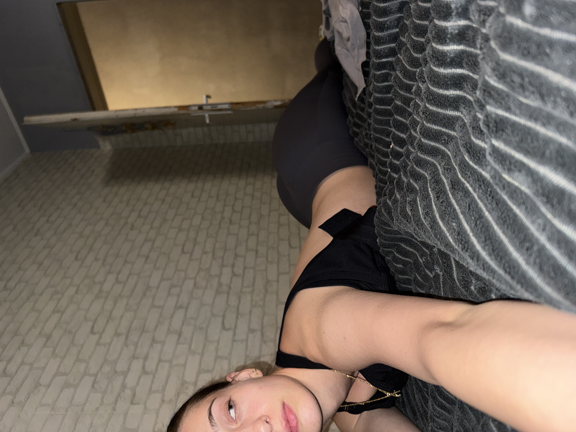 The image depicts a person lying down, possibly on a bed or couch, with their head towards the bottom of the frame. The background includes a wall with a brick-like pattern and a partially visible ceiling. The person is wearing what appears to be a black top and gray shorts. Part of a striped blanket or towel is visible on the right side of the frame. It is not possible to determine if the scene is in Sofia, Bulgaria, or any specific location without additional information.