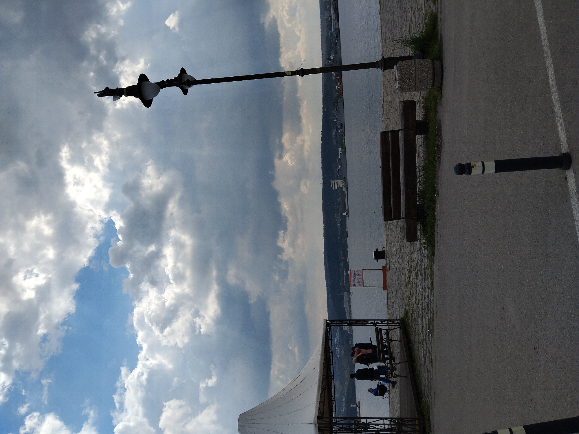 The image shows a scene on the Varna Sea Garden promenade in Varna, Bulgaria. There's a view of the Black Sea on the right, a grey paved walkway, and the sky with scattered clouds on the left. A dark-colored lamppost with two lamps is visible, reaching from the right toward the sky. Next to the walkway are a wooden bench, a bollard, and a waste receptacle. A small structure with a white awning and black metal supports stands near the edge of the promenade. Some people are sitting and walking under the awning. The background shows the coastline.