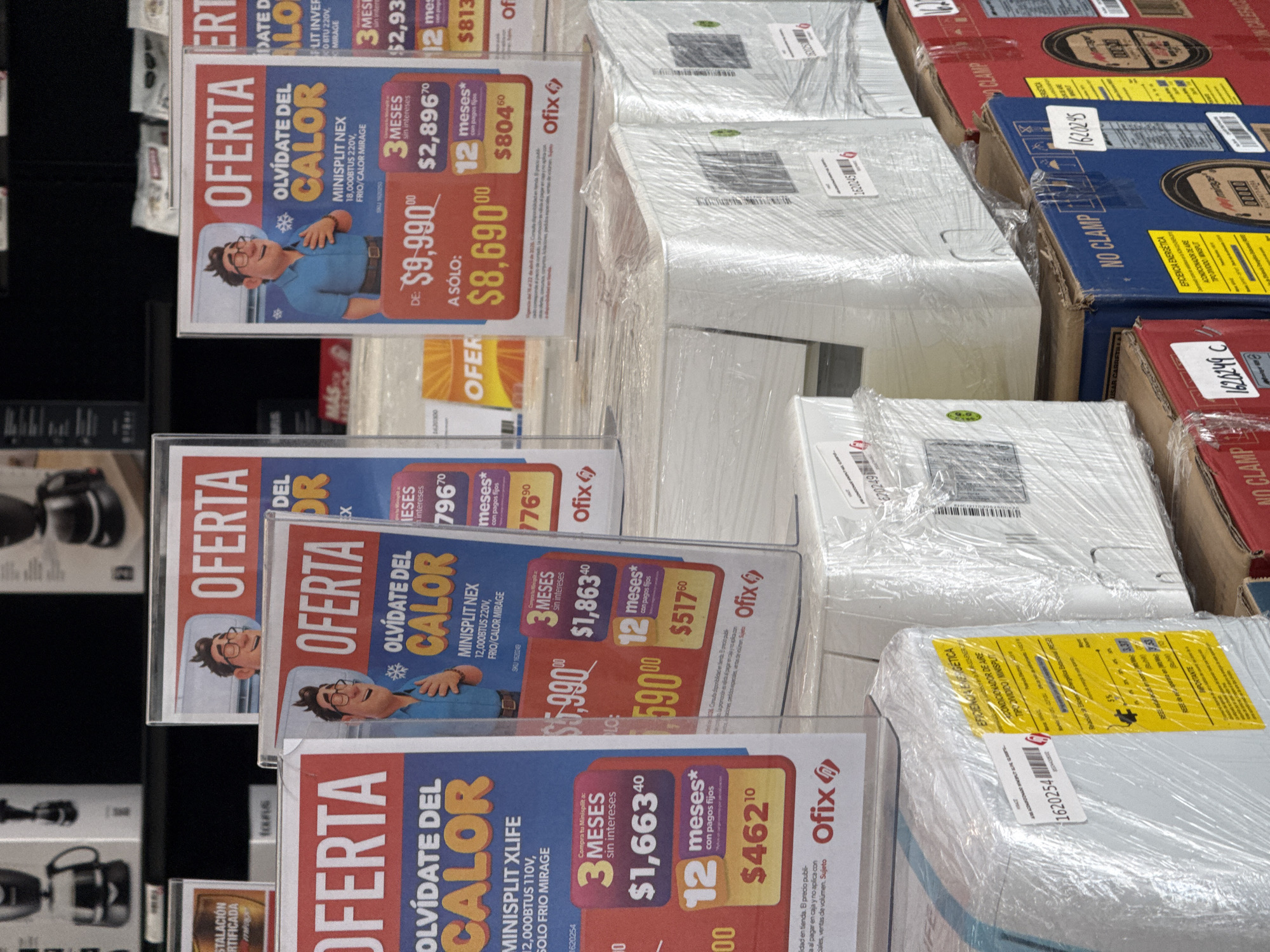 The image captures a retail display, likely within a store in San Bernardino Tlaxcalancingo, Mexico, showcasing multiple air conditioning units. The scene is dominated by stacked cardboard boxes of various sizes, many wrapped in clear plastic film, signifying new merchandise. Several prominent "OFERTA" (Offer) signs are displayed vertically, promoting "MINISPLIT" air conditioners.
Each sign features a cartoon man looking relaxed and content, with text emphasizing "OLVÍDATE DEL CALOR" (Forget the heat). The promotions include models like "MINISPLIT NEX 18,000BTUS 220V, FRIO/CALOR MIRAGE" initially priced at $9,990.00, now "A SÓLO: $8,690.00", with payment options of $2,896.70 over 3 months or $804.60 over 12 months. Another sign details a "MINISPLIT NEX 12,000BTUS 220V" for $5,990.00, offering 3 months at $1,863.40 or 12 months at $517.60. A "MINISPLIT XLIFE 12,000BTUS 110V, SOLO FRIO MIRAGE" is also advertised with payment plans of $1,663.40 for 3 months or $462.10 for 12 months. The brand "Ofix" is visible on the signs.
The boxes have various labels, including product numbers like "1620249" and "1620254", and logistical instructions such as "NO CLAMP" and "USAR CARRETILLA" (use forklift). A yellow label on one box mentions "EFICIENCIA ENERGETICA ACONDICIONADOR DE AIRE TIPO DIVIDIDO / MINISPLIT" (Energy Efficiency Air Conditioner Split Type / Mini-Split). The overall impression is one of a product launch or seasonal sale for cooling appliances.