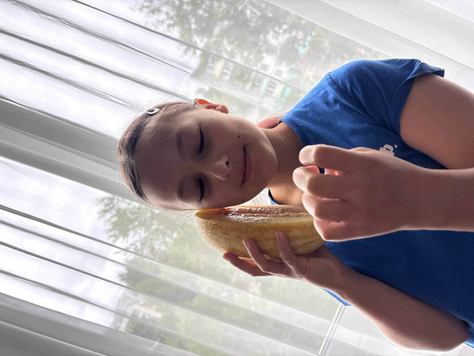 A young girl, likely between 8 and 12 years old, stands in front of a white sheer curtain indoors. She has medium-length brown hair with pink tips, pulled back and secured with a silver clip. She is wearing a blue t-shirt. Her eyes are closed, and she is holding a slice of melon to her right ear as if it were a phone, with her left hand supporting it and her right hand near her face. The background visible through the curtain suggests an urban environment, possibly in Chaikovsky, Russia, with the blurry shapes of buildings and trees visible. The lighting is soft and diffused, coming from the window behind her.