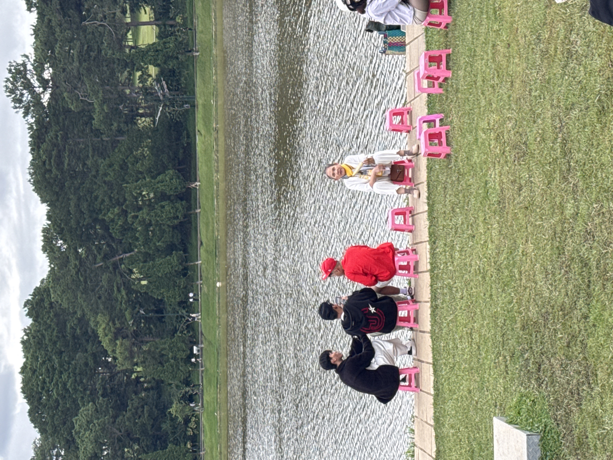 Here is a description of the image:
A medium shot shows a group of people sitting on bright pink plastic stools along the concrete edge of a body of water. The water appears to be rippled. The first person in the row is a woman wearing light-colored clothing and a yellow scarf. She is smiling. The next person is wearing a red shirt and hat. Two more people are wearing black jackets. There is an extra stool between each person. Behind them is a grassy area. Beyond that is a line of trees. The sky is overcast.
This scene is likely located on the shore of Xuan Huong Lake in Da Lat, Vietnam, based on its appearance and typical tourist activities.