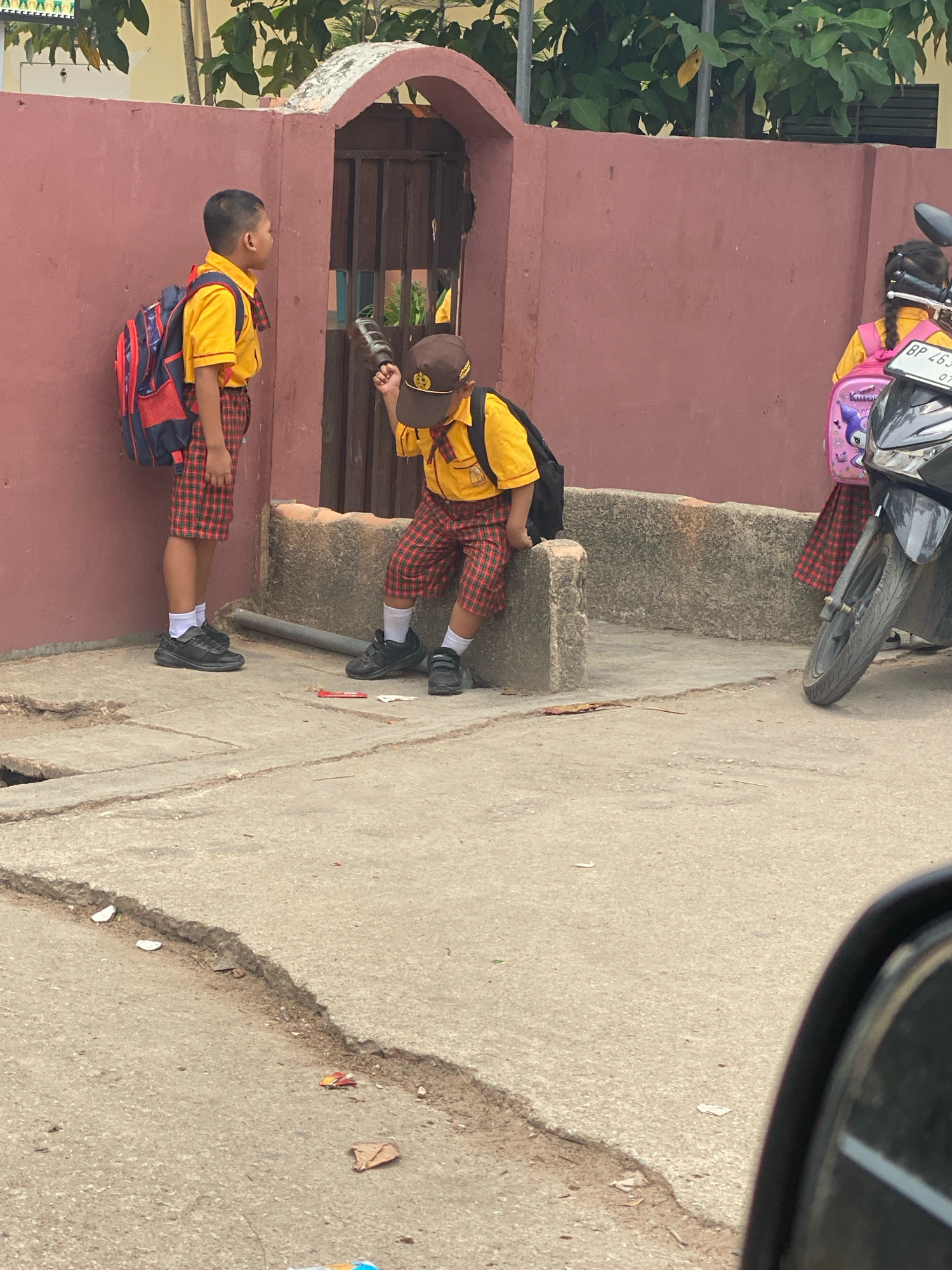 In Batam, Indonesia, two young school children are seen in front of a reddish-brown wall. The child on the left stands wearing a yellow shirt, plaid shorts, and a backpack, looking to the right. The child on the right is seated and wearing a similar uniform, holding a dark object, possibly a toy or a tool, and looking towards the child on the left. Another child, partially visible, is seen on the right, wearing a pink backpack and a yellow shirt, standing next to a motorcycle. The ground in front of the wall is concrete with a prominent crack. The environment suggests an outdoor setting, possibly near a school or residential area, given the presence of children in school uniforms.