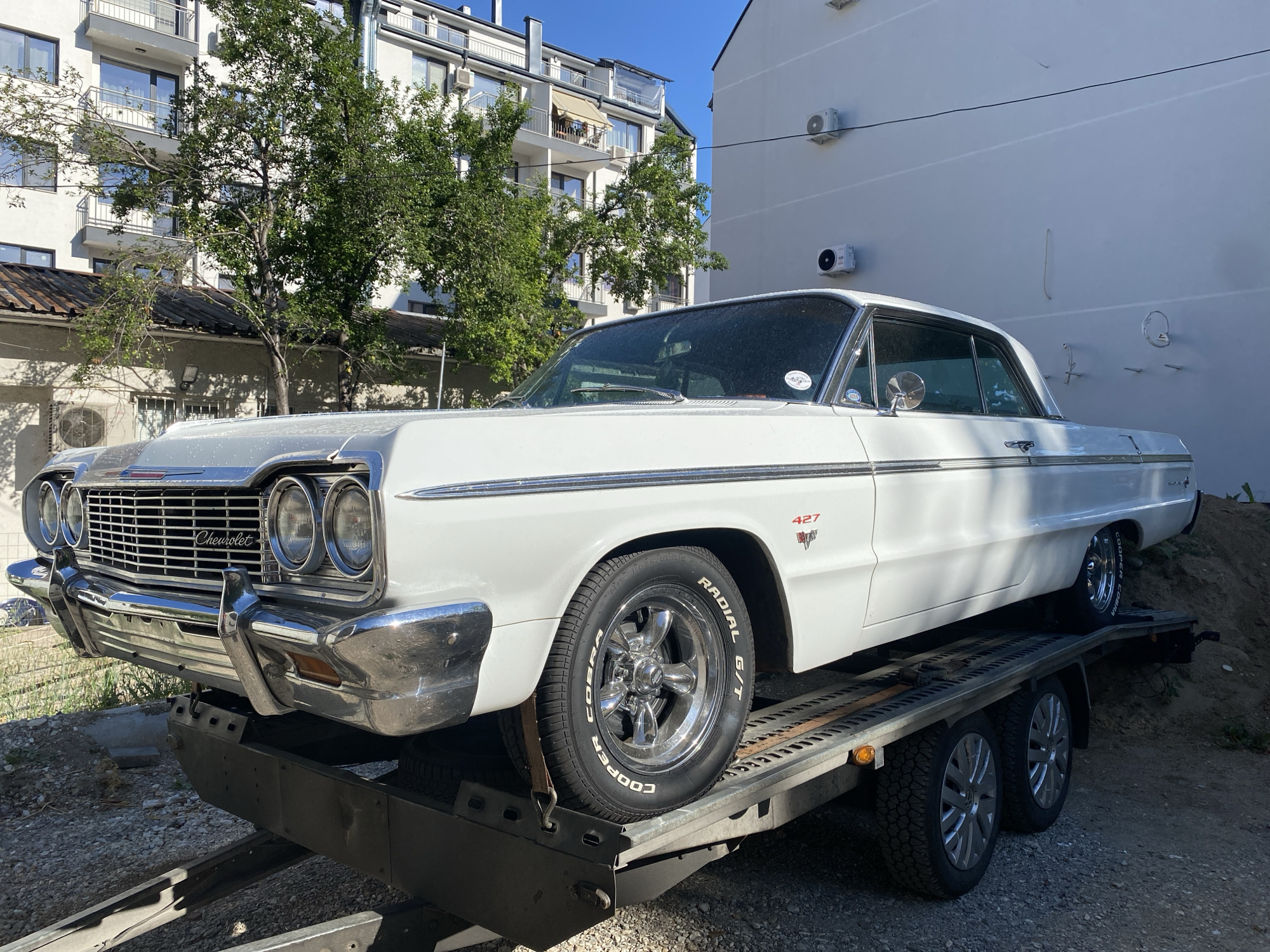 A white Chevrolet Impala 427 is parked on a car trailer, facing right. The car is a classic two-door coupe with chrome detailing, prominent headlights, and a large grille. The trailer is loaded onto a gravel surface, with a steep dirt embankment visible behind it. In the background, a multi-story white building with balconies and trees is seen under a clear blue sky. The scene appears to be an urban environment, specifically in the city of Pleven, Bulgaria, suggested by the architectural style and surrounding elements. No people are visible in the image. The car is the primary subject, with the trailer and the urban background providing context.