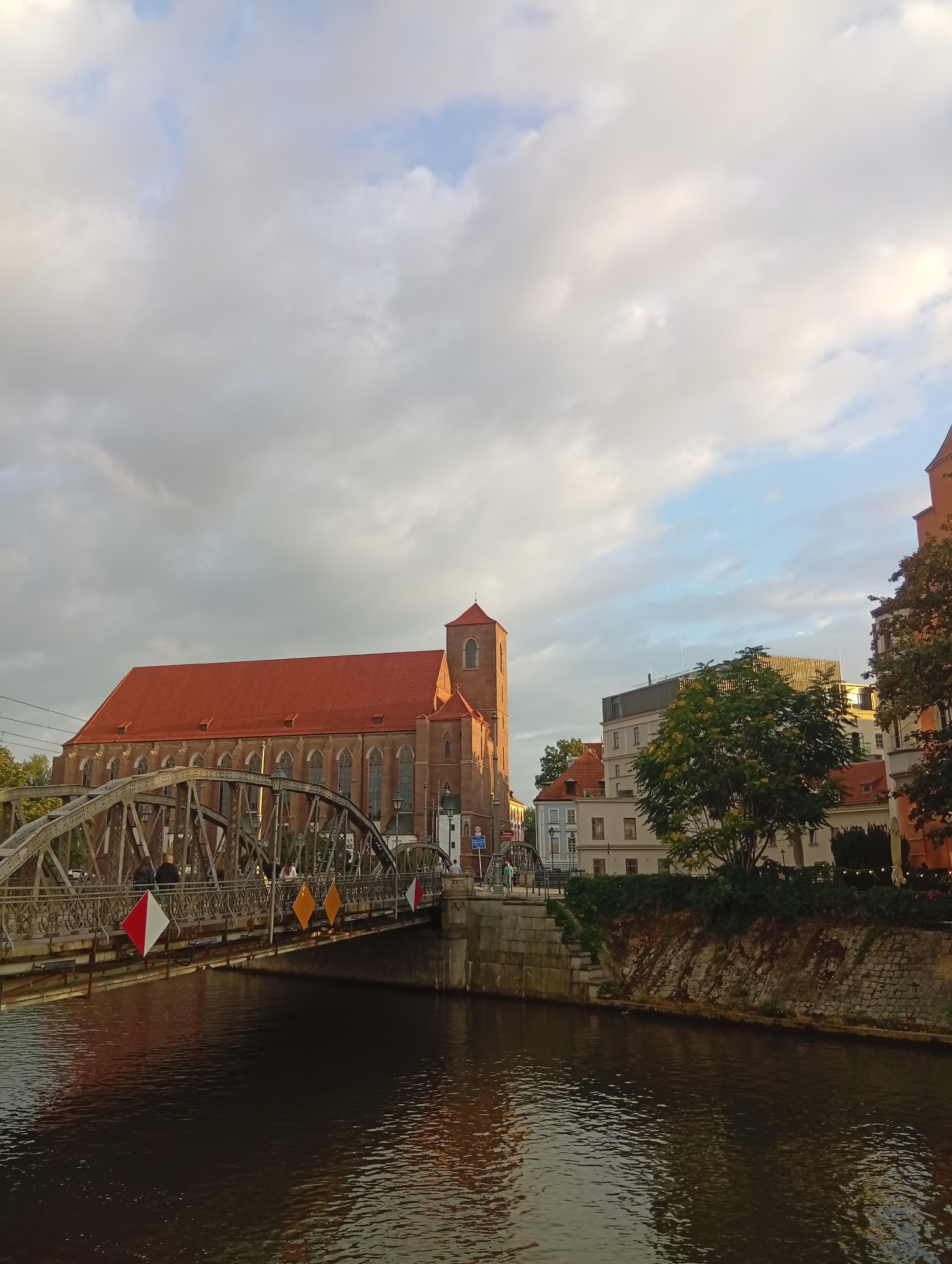The image captures a view of the Oder River in Wrocław, Poland, with the Cathedral of St. John the Baptist visible on the left bank. A metal arch bridge spans the river, connecting to the cathedral's vicinity. The cathedral itself is a large, brick building with a prominent red tiled roof and a tall, slender tower topped with a spire. The sky is filled with a mixture of white and grey clouds, with patches of blue visible. The water in the river reflects the light from the sky and the surrounding buildings. Several people are walking on the bridge, some of whom are indistinct. The bridge is decorated with diamond-shaped flags in red and white and yellow colors. The riverbank on the right is lined with stone walls and lush green vegetation, with several buildings visible behind the trees. The overall scene suggests a late afternoon or early evening setting, with the warm sunlight illuminating parts of the cathedral and the surrounding architecture.