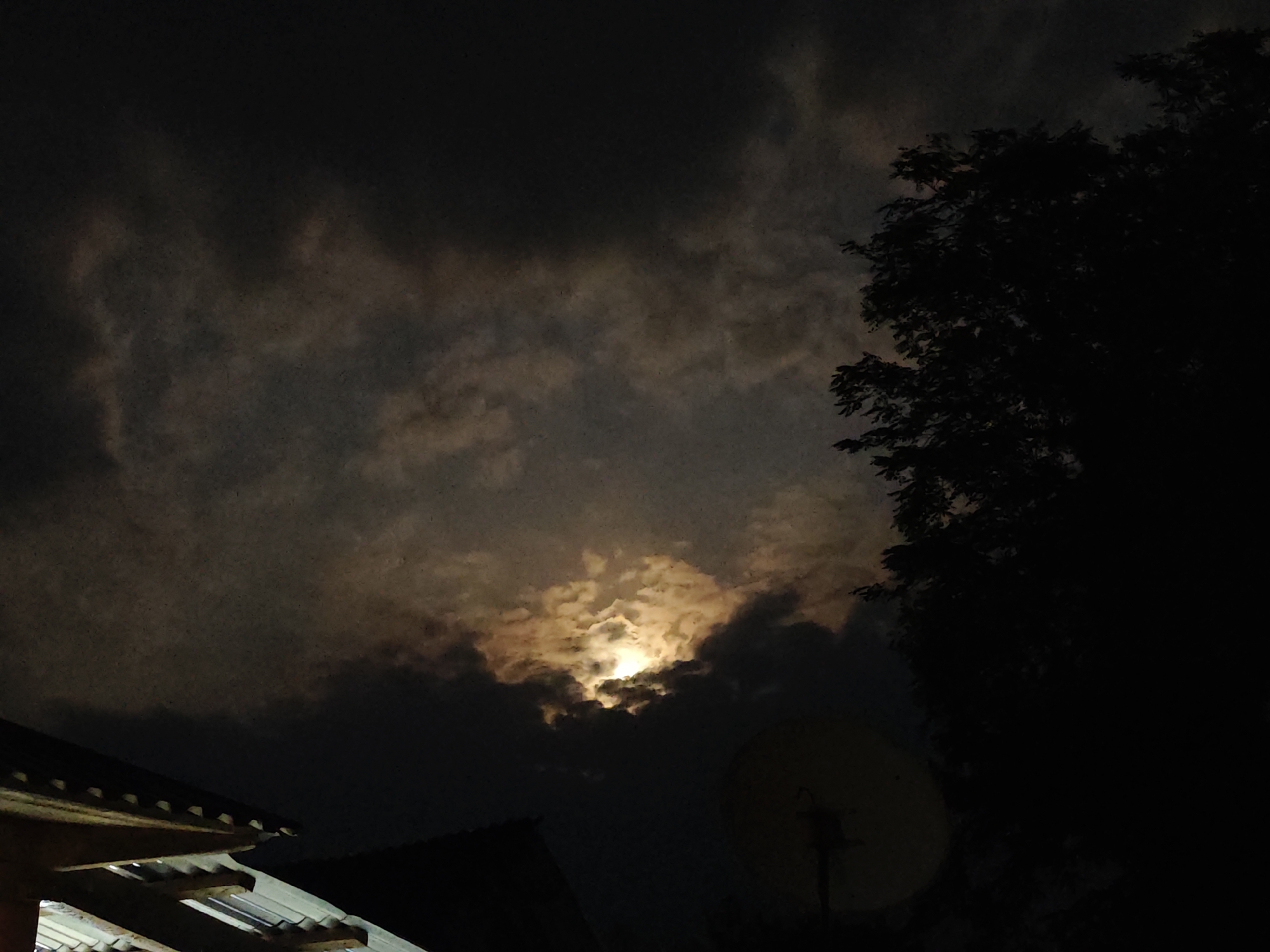 The image depicts a nighttime scene dominated by a cloudy sky with the moon partially visible and illuminating the clouds with a warm, yellow glow. In the foreground, the dark silhouette of a tree with dense foliage occupies the right side of the frame. To the left, the angled roof of a building, possibly a house, is visible with some structural details of its underside. Further to the right and lower down, a satellite dish on a stand is partially visible in the dark. The overall environment suggests an outdoor setting at night, likely in a residential area given the presence of the building and satellite dish. The location is identified as Бчара ақыҭа ахадара, Abkhazia. There are no people or visible actions depicted.
