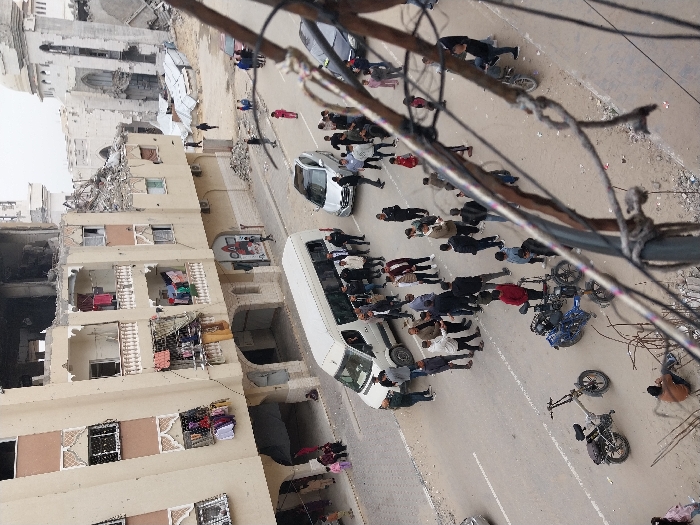 A chaotic scene unfolds on a street next to damaged buildings. A white bus and a white car are positioned among a crowd of people, many dressed in dark attire. Several motorcycles lie scattered on the ground, some overturned. Power lines run across the foreground, partially obscuring the view. The buildings show signs of destruction with exposed interiors and debris. The overall atmosphere conveys distress and disarray.