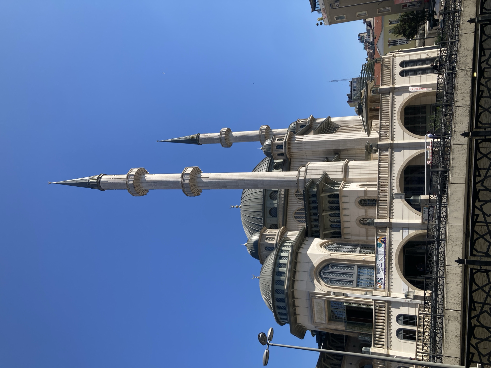 The image displays a large, ornate mosque with two prominent minarets against a clear blue sky. The architecture features light-colored stone, multiple domes, and arched windows. A black metal railing runs along the foreground, partially obscuring the lower part of the building. Several people are visible on balconies and near an entrance, suggesting the mosque is open to visitors or congregants. Banners with Turkish text are hung on the building's facade, including one that reads "YAZ OKULU" which translates to "Summer School". The bright sunlight and deep blue sky indicate it is a sunny day, likely during daylight hours. The location is identified as Beyoğlu, Turkey. The overall scene conveys a sense of grandeur and cultural significance.