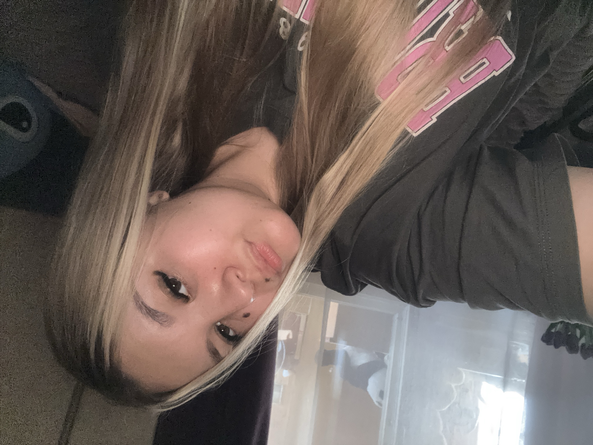 A young woman with blonde hair and a dark t-shirt is lying down and taking a selfie. She has a beauty mark on her cheek and wears dramatic eyeliner. The background shows a window with diffused light and some blurry objects. She is indoors, likely in a residential setting, with a dark sofa or blanket visible. No specific location in Bolshoy Bukor, Russia can be determined from the image.