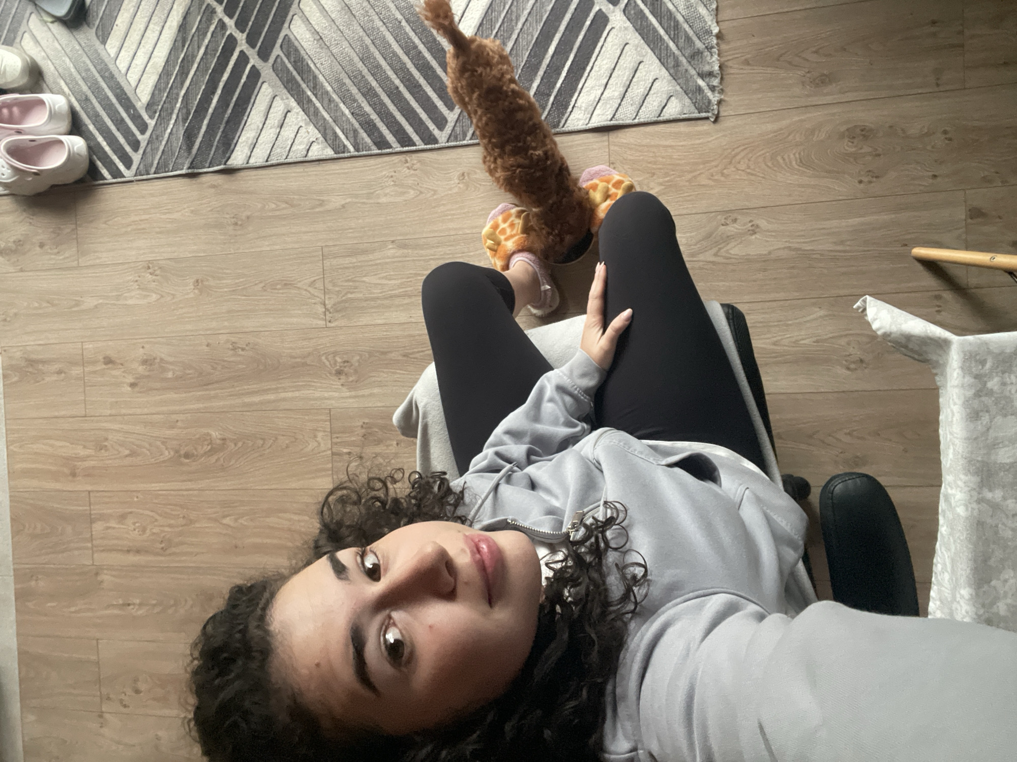 The image captures an indoor scene from a high-angle perspective, likely a selfie taken by the person depicted. The main subject is a young woman with dark, curly hair, lying or reclining on a black chair. She is wearing a light grey zipped hoodie and black leggings, looking directly at the camera with a relaxed expression and a slight smile. Her left hand rests on her lap.
Between her feet, a small, fluffy brown dog with a long tail is visible, appearing to interact with or chew on one of her orange and yellow animal-themed slippers. Another pair of white slip-on shoes with pink lining is seen near a patterned grey and white rug in the upper left corner of the frame. The room features light-colored wooden floorboards throughout.
Other objects in the background include parts of an armchair with wooden legs on the right side. The overall setting suggests a comfortable, domestic environment, possibly a living room or bedroom. The lighting indicates it is daytime, but no specific light sources or windows are visible. There is no visible text in the image. While the provided context suggests Sofia, Bulgaria, there are no specific visual cues within the image to confirm this location.