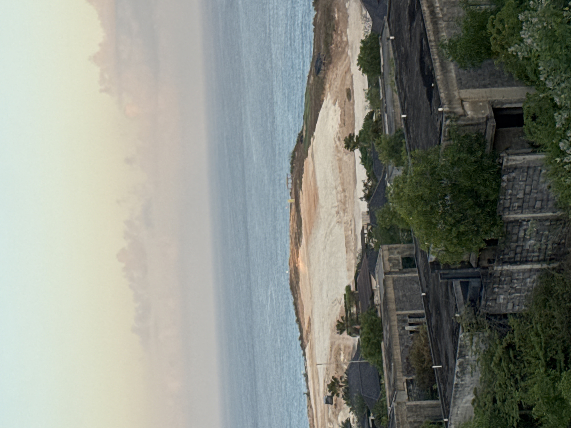 The image depicts an elevated, long-shot view of a landscape bordering the ocean. In the foreground, several weathered, low-rise buildings with flat, dark roofs are visible. These structures appear to be constructed from concrete blocks and show signs of age and disrepair, with some partially obscured by lush green foliage. Beyond these buildings, a large, uneven expanse of light-colored earth dominates the mid-ground, likely a construction or development site, with a few small structures and scattered vegetation on its surface. In the distance, the vast expanse of the ocean stretches to the horizon, meeting a softly colored sky with pale clouds, suggesting either dawn or dusk. The scene is located in Kuta Selatan, Indonesia, a region known for its coastal beauty and ongoing development.