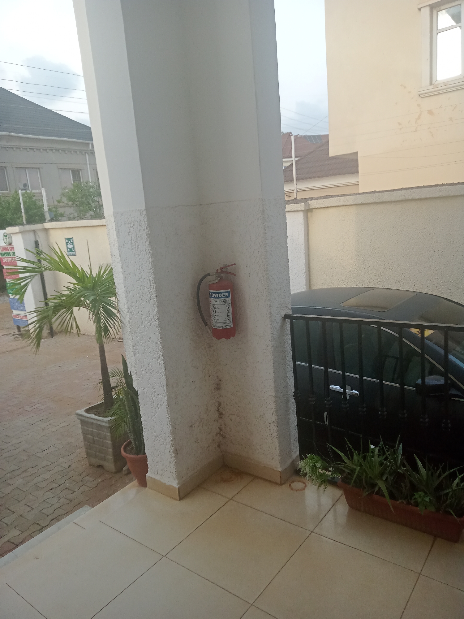 The image shows the corner of a building in what appears to be an urban area, possibly Gwarinpa, Nigeria, given the prompt's instruction. A thick, white support column dominates the foreground, with a red fire extinguisher mounted on its wall. Adjacent to the column are various potted plants, with a black car parked behind a black metal fence. Beyond the column and plants, there is a view of other buildings, including a grey building with windows and a beige building with a window and a wall. The ground is tiled with beige tiles.