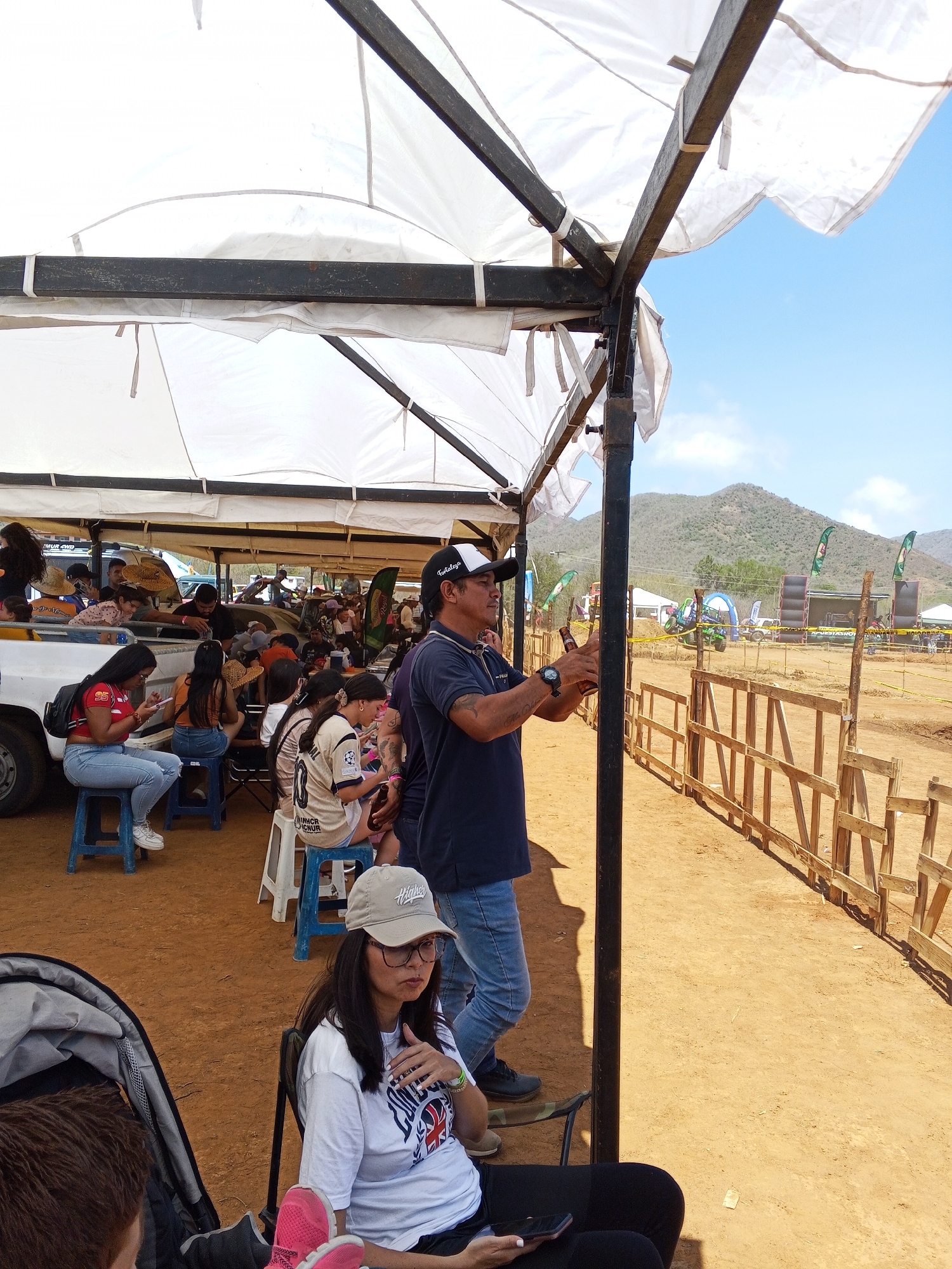 The image captures a scene at an outdoor event in Unknown, Unknown. Under a white tent structure, people are seated on stools and chairs. Some are focused on their phones, while others seem to be watching the event. A man in a baseball cap, navy t-shirt, and jeans stands near a support beam, holding a bottle and looking towards a fenced-off area. The background features a dirt ground, wooden fencing, and a hill in the distance. The scene suggests a casual and observational atmosphere at a local gathering or event.