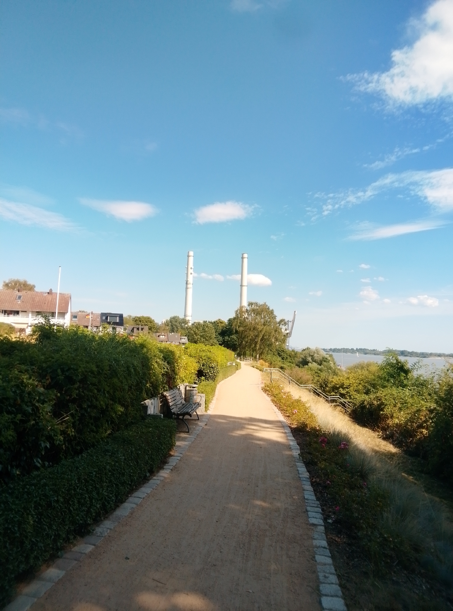 A paved pathway leads through a park area in Wedel, Germany, on a sunny day with a clear blue sky and scattered white clouds. On the left side of the pathway, a meticulously trimmed green hedge borders the path, with a wooden bench and a trash can visible amongst the foliage. Beyond the hedge, houses with tiled roofs are visible. On the right side of the pathway, the terrain slopes downwards towards a body of water, with lush green trees and bushes lining the bank. In the distance, two tall, white industrial chimneys stand prominently against the sky. A large crane is also visible in the background, suggesting an industrial or port area nearby. The overall scene depicts a serene and well-maintained public space.