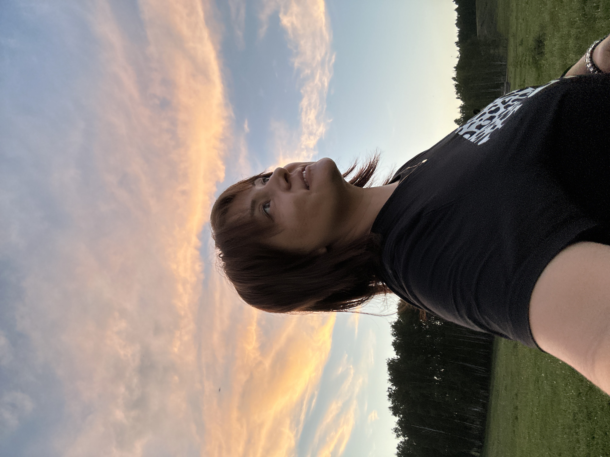 An outdoor, medium shot captures a woman facing right, looking up at a dramatic sunset. Her head and shoulders are in profile, with her brown hair framing her face. She wears a black t-shirt with a white pattern on the left sleeve. The background is a vast, open field with a line of dark trees in the distance, beneath a sky painted with hues of pink, orange, and blue from the setting sun. The image is a selfie, with the photographer's arm and hand visible in the lower left corner.
