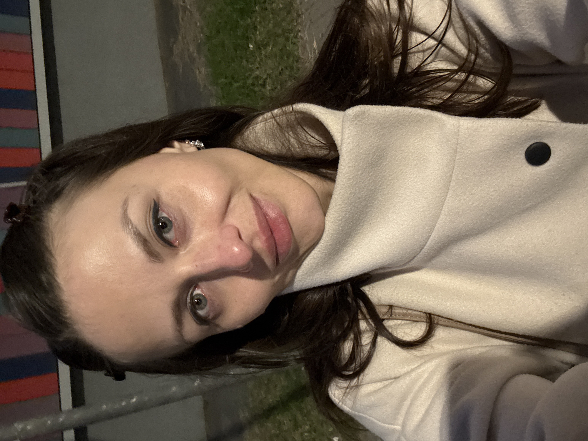 A woman with light skin, brown hair, and grey eyes is centered in the frame, angled slightly towards the viewer. She is wearing a light-colored coat with a visible black button. A small, decorative earring is visible in her left ear.
Behind her, a blurry background suggests an outdoor setting. In the upper portion, there is greenery. To her left is a structure with horizontal stripes of varying colors. A metal railing is visible in the lower-left corner.
The location is Saint Petersburg, Russia.