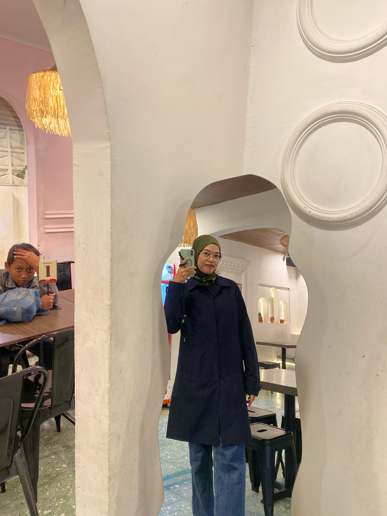 The image captures an indoor scene, possibly a cafe or restaurant, with distinct architectural features. In the foreground, a young person with their hand on their forehead sits at a table with a number "1" sign. They are wearing a denim jacket and have a blue backpack beside them. To the right, a person wearing a hijab, glasses, a dark coat, and blue jeans is taking a mirror selfie with a smartphone. The background reveals white walls with decorative circular moldings and arched doorways. Behind the person taking the selfie, tables and stools are visible, along with a wall with arched niches containing decorative objects. A yellow, tasseled light fixture hangs from an arched ceiling. The overall ambiance suggests a modern, stylized interior.