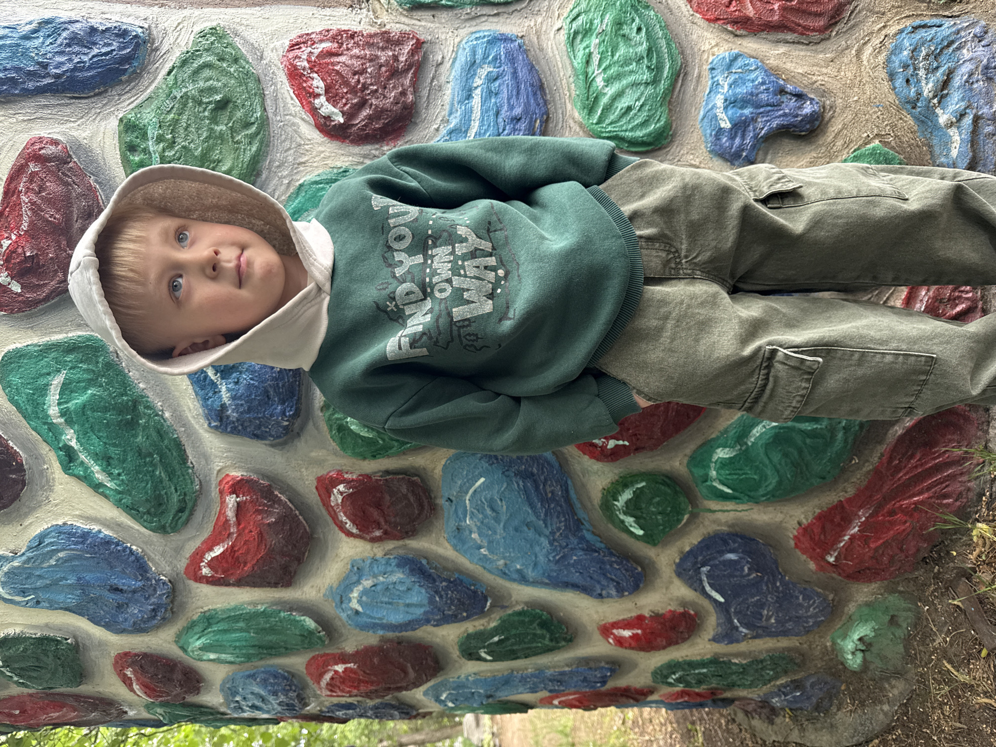 A young child with blond hair and blue eyes is lying on a structure resembling a wall made of painted stones. The child is wearing a hooded sweatshirt, with the hood up, and green pants. The sweatshirt has the words "FIND YOUR OWN WAY" printed on it. The stones are painted in a variety of colors including red, blue, and green. The structure is outdoors. According to metadata, the image was taken in the city of Chaikovsky, Russia.