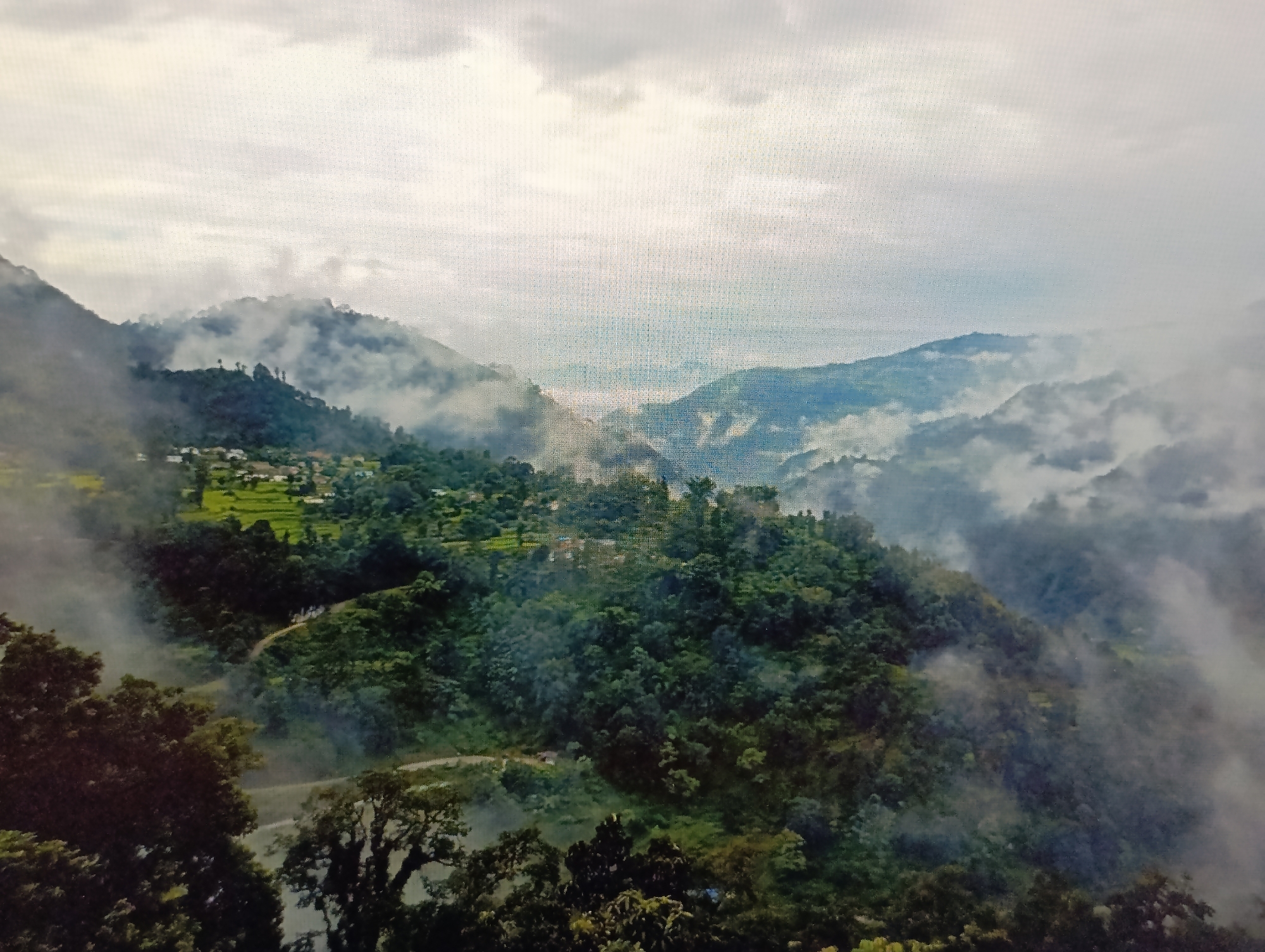 The image depicts a misty mountain landscape, with rolling green hills covered in dense vegetation. A small village is nestled on one of the slopes, with buildings visible amidst the trees and terraced fields. A winding road snakes its way through the terrain. The sky is overcast with clouds, adding to the atmospheric and somewhat mysterious feel of the scene. There is no indication of the city of Dubăsari town council, Moldova within the image.