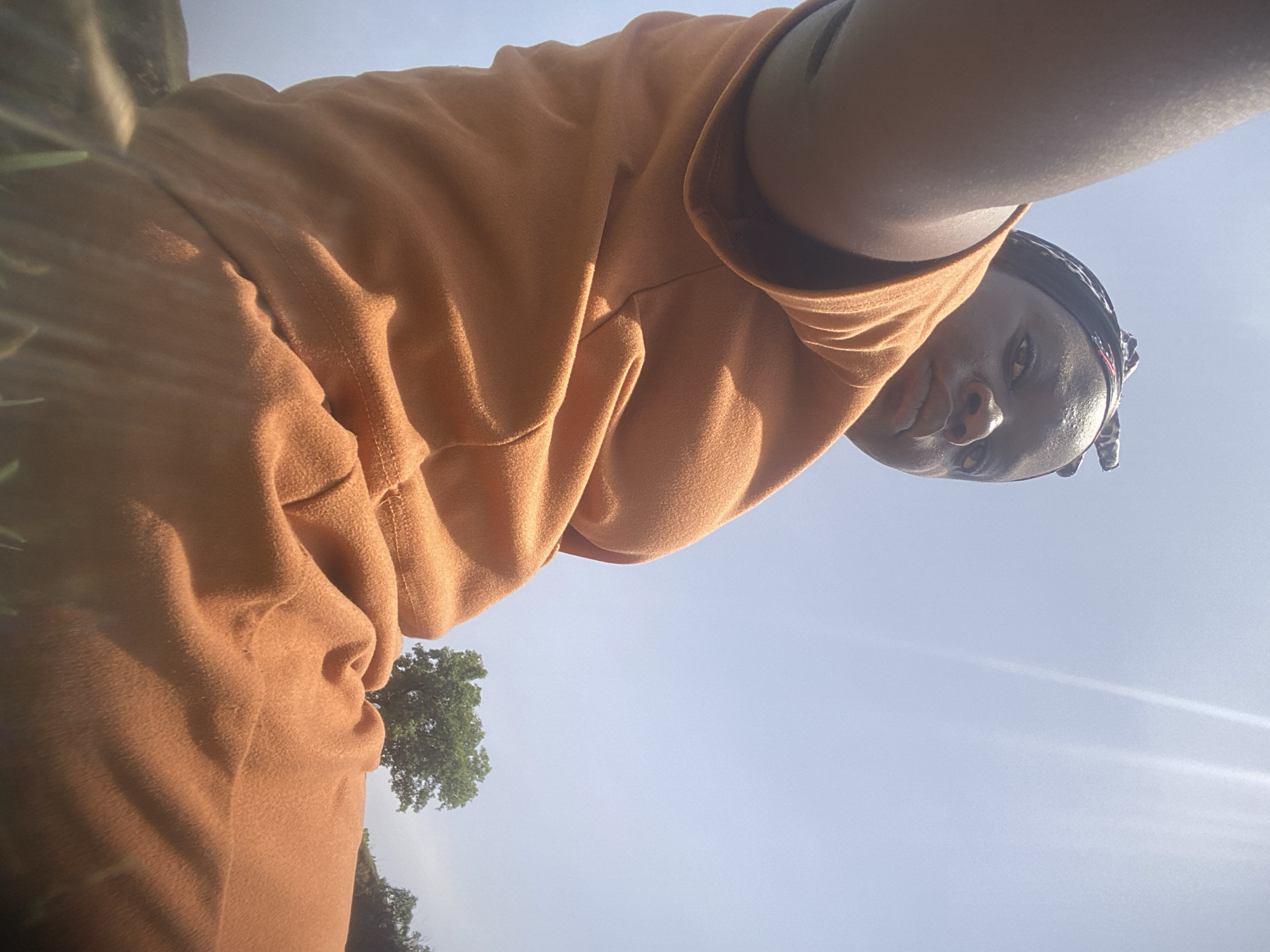 The image, taken in Kukum, Nigeria, presents a low-angle perspective of a person with dark skin looking directly into the camera. The individual is positioned against a bright, clear blue sky, suggesting an outdoor setting under strong sunlight.
The subject, likely female, wears a loose-fitting, long-sleeved top and what appears to be matching bottoms in a warm orange-brown color. The fabric exhibits a soft, slightly textured quality, possibly fleece or thick cotton, with visible seams and folds indicating a relaxed fit. A dark, patterned head covering or bandana is worn around the head. The subject's face is angled slightly downwards towards the camera, displaying a calm, neutral, or subtly pensive expression. Highlights on the skin are noticeable, consistent with direct sunlight.
In the background, a portion of a lush green tree with dense foliage is visible in the lower part of the frame (when the image is viewed upright). The vast expanse of clear blue sky fills most of the background, with some soft light flares indicative of bright daylight. The scene captures a casual, sunlit moment outdoors. No text is visible in the image.