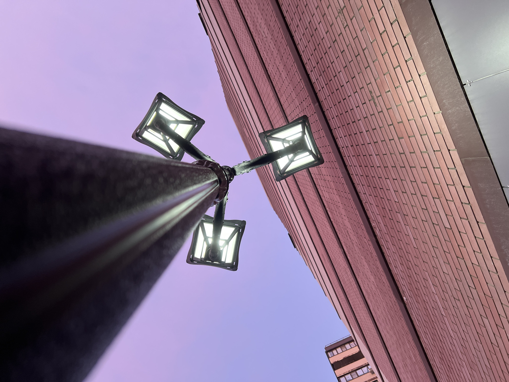 The image shows a low-angle view of a streetlamp with three illuminated square lanterns against a twilight sky. Adjacent to the lamp is a tall brick building. A portion of another building is visible in the lower right corner. The scene appears to be urban. There is no evidence in this image to suggest it is in the city of Krasnodar, Russia. No people, actions, or interactions are depicted.