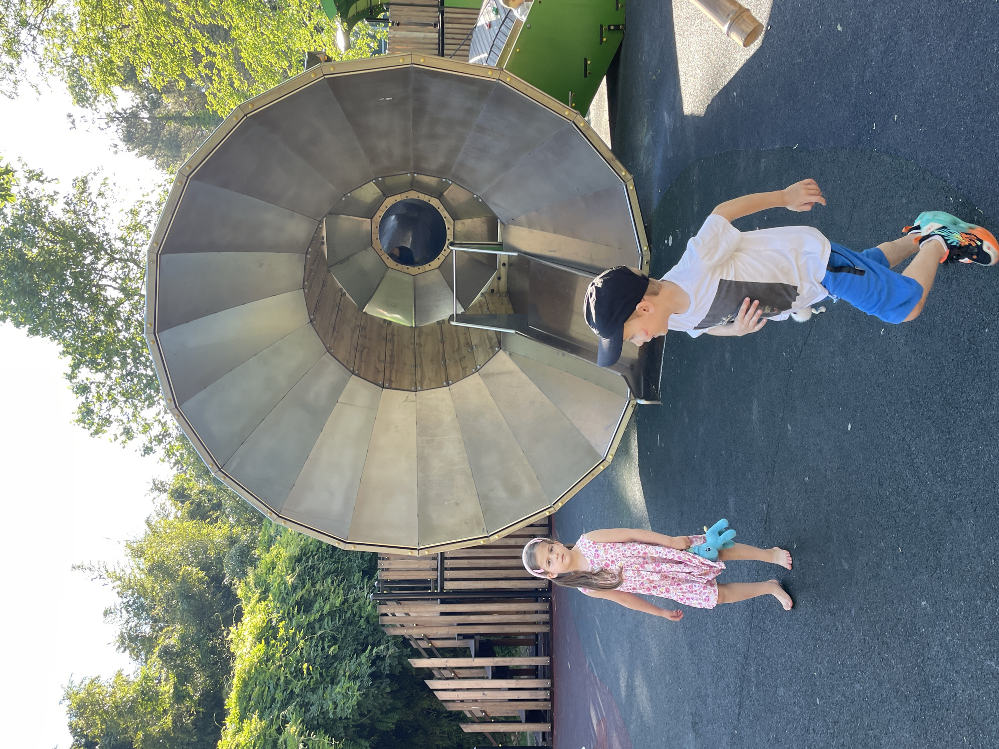 Two children are on a playground. A girl in a pink floral dress is standing on the left, barefoot, holding a blue toy. A boy in a white t-shirt and blue shorts is running on the right, wearing a black baseball cap and colorful sneakers. Behind them is a large, metallic, funnel-shaped play structure with a slide. In the background, there is a wooden climbing structure and lush green trees. The ground is a dark, rubberized surface. The scene appears to be outdoors on a sunny day. There is no indication that this image is located in the city of Sochi, Russia.