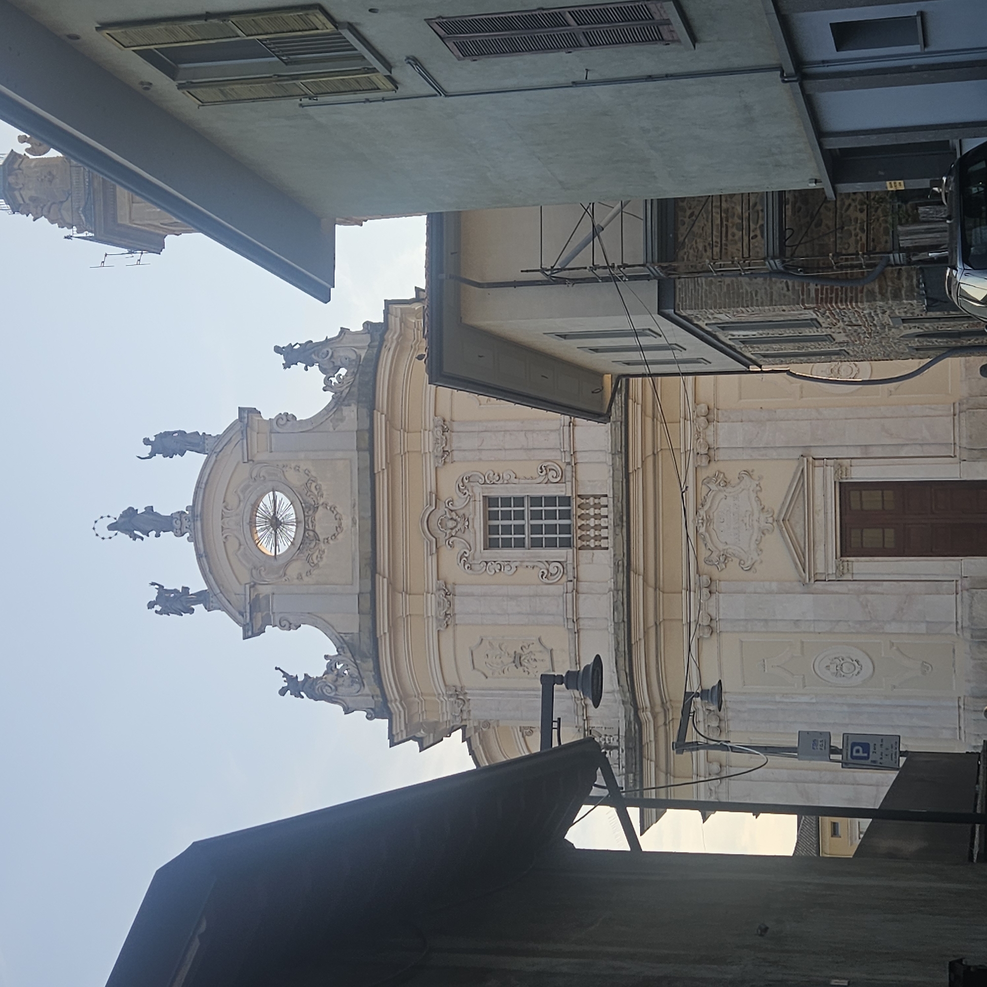 The image shows the facade of a baroque-style church in Calcinate, Italy. The church has a cream-colored exterior with ornate architectural details, including sculptures of figures at the top and a large clock face. In front of the church, there are buildings that partially obscure the view. One building on the right has light-colored walls and brown shutters, while another on the left is dark and appears to be closer to the viewer. A street lamp with two lights is visible on the left side, and parking signs are mounted on its pole. The sky is clear and a pale blue. The lower part of the church features a large wooden door framed by columns.