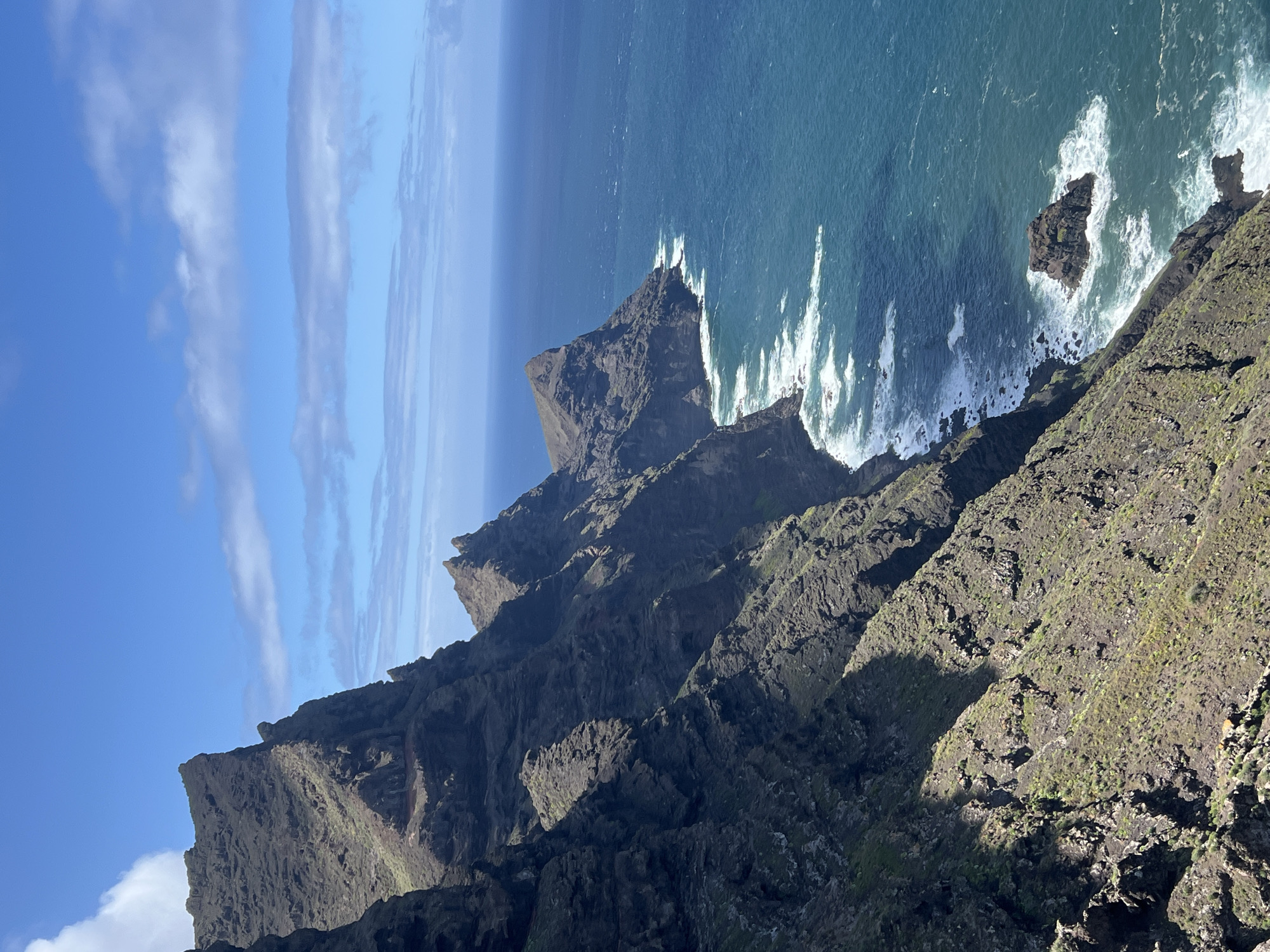 The image shows a mountainous coastal landscape near Tirma, Spain. Steep, rocky cliffs plunge into the Atlantic Ocean, where waves crash against the shore. The sky is blue with scattered clouds. There are no people visible in the shot. The scene emphasizes the rugged terrain and the interplay between the land and the sea.