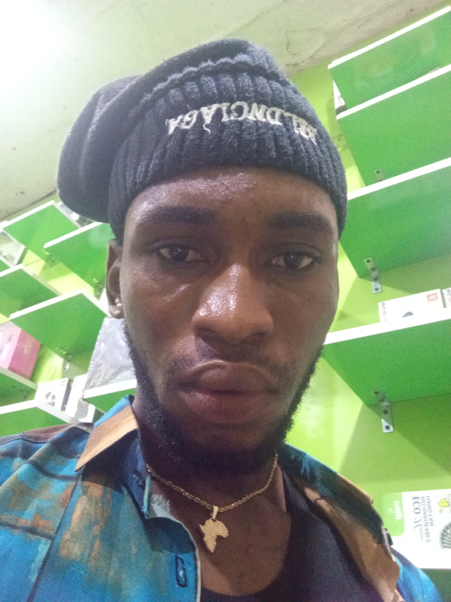 A young Black male is shown in a close-up, head-and-shoulders selfie. He is wearing a black knit cap with white lettering, a blue and brown patterned shirt, and a gold chain with an Africa-shaped pendant. He has a beard and appears to be in an indoor setting with green shelves in the background. The presence of shelves stocked with various items suggests a retail or storage environment. The specific location in Lokoja, Nigeria, cannot be determined from the visual information alone.