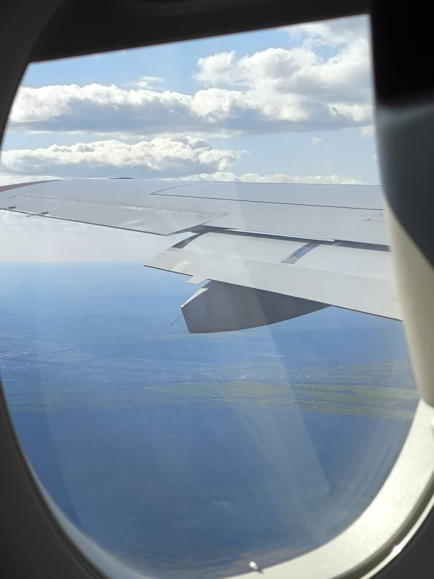 An aerial view from an airplane window captures a wide landscape under a partly cloudy sky. The lower-left and right edges of the image show the gray-white curved frame of an aircraft window. The main subject is the light gray wing of an airplane, extending from the left of the frame diagonally towards the upper right, with visible control surfaces and a structural component (possibly a fairing or pylon) beneath its outer section. Beyond the wing, the scene is divided horizontally: the upper half depicts a bright blue sky filled with scattered, fluffy white cumulus clouds, arranged in several horizontal layers. The lower half of the background reveals a vast, serene body of blue water, likely a large lake or sea, which dominates the mid-ground below the wing. Adjacent to and beyond the water, patches of green land are visible, featuring dense vegetation and faint indications of human development, such as dispersed structures or roads, in the distance. The lighting suggests daylight conditions, with sunlight reflecting off the wing and the water surface. No individuals are present in the image.