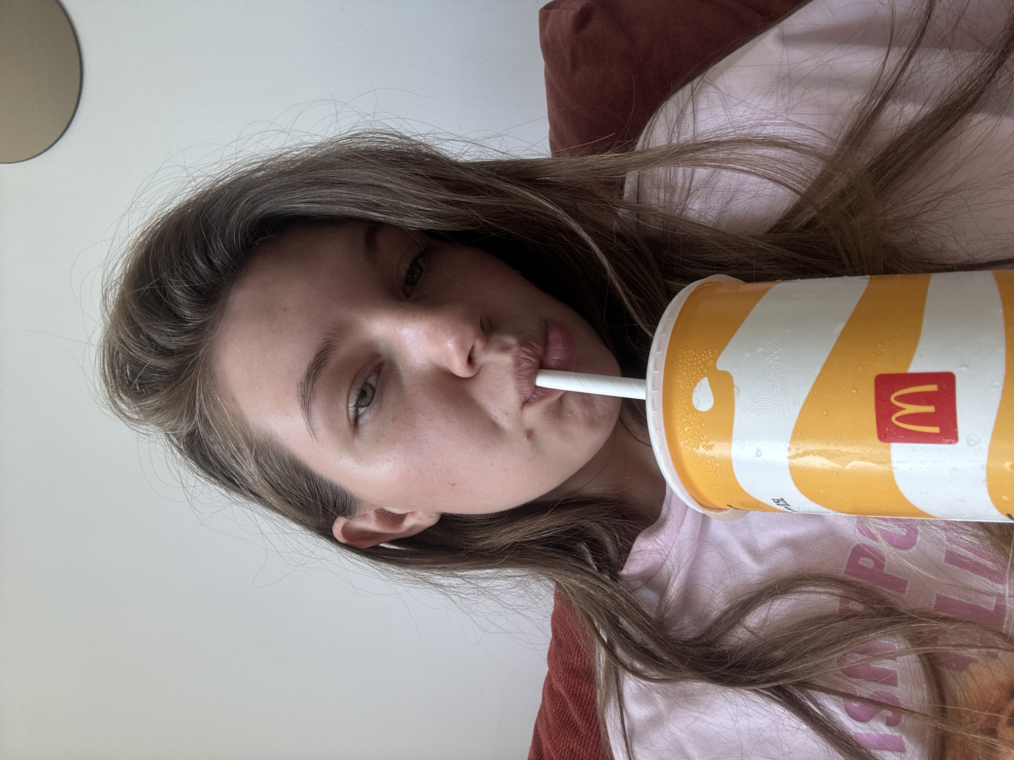 A young woman with light brown hair is lying down and drinking from a large McDonald's cup with a straw. The cup is primarily white and yellow with the McDonald's logo. The woman is wearing a light pink shirt. The background includes a light-colored wall, a brown cushion, and a portion of a light-colored object. Location information is not discernible from the provided image.