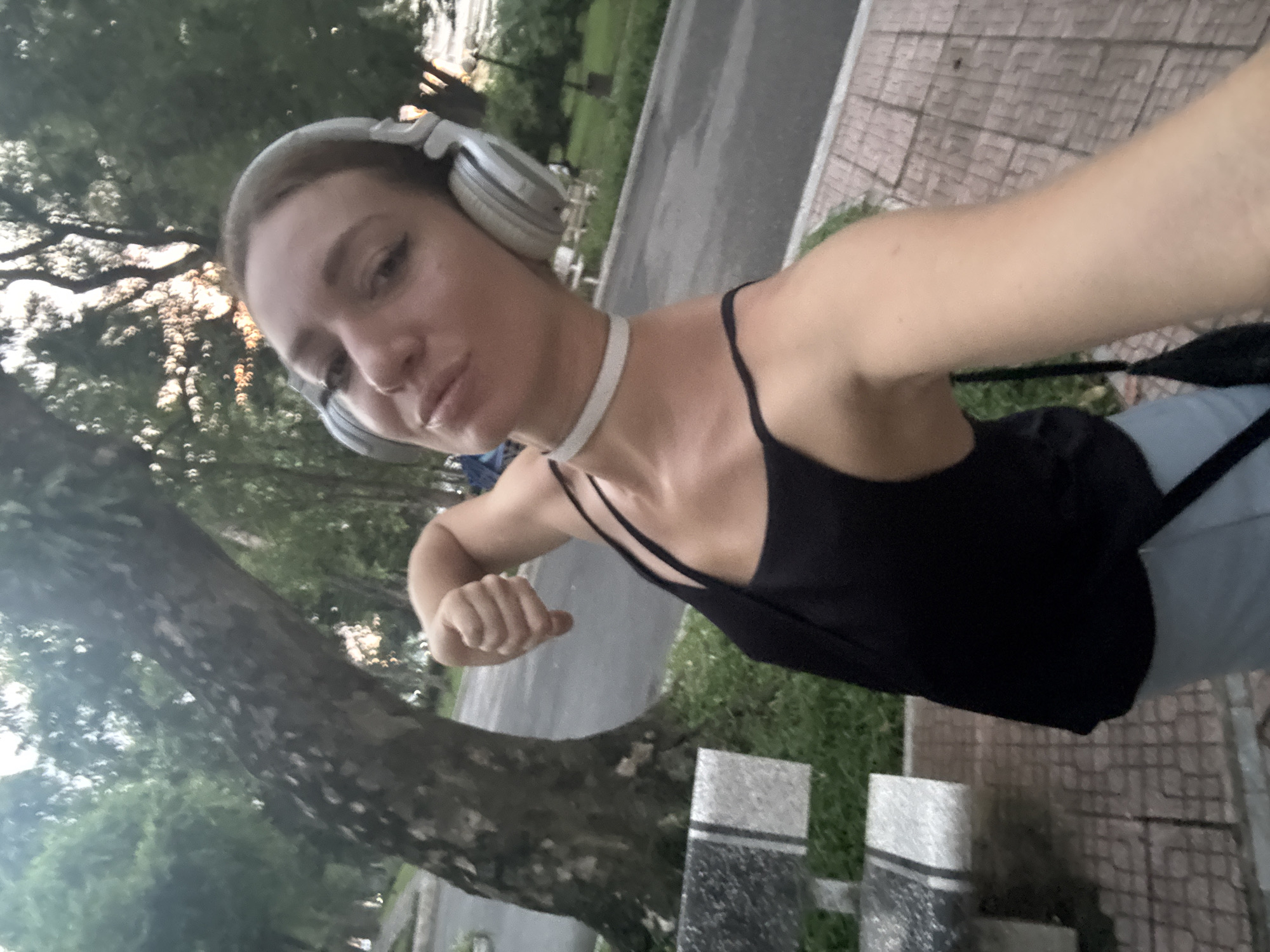 In Hà Nội, Vietnam, a young woman with blonde hair is taking a selfie in a park. She is wearing a black tank top, light blue pants, a white choker, and silver headphones. She is flexing her arm and puckering her lips. Behind her, a large tree with textured bark is visible, along with park benches, a paved walkway, and lush green foliage. The overall scene suggests an outdoor recreational setting.