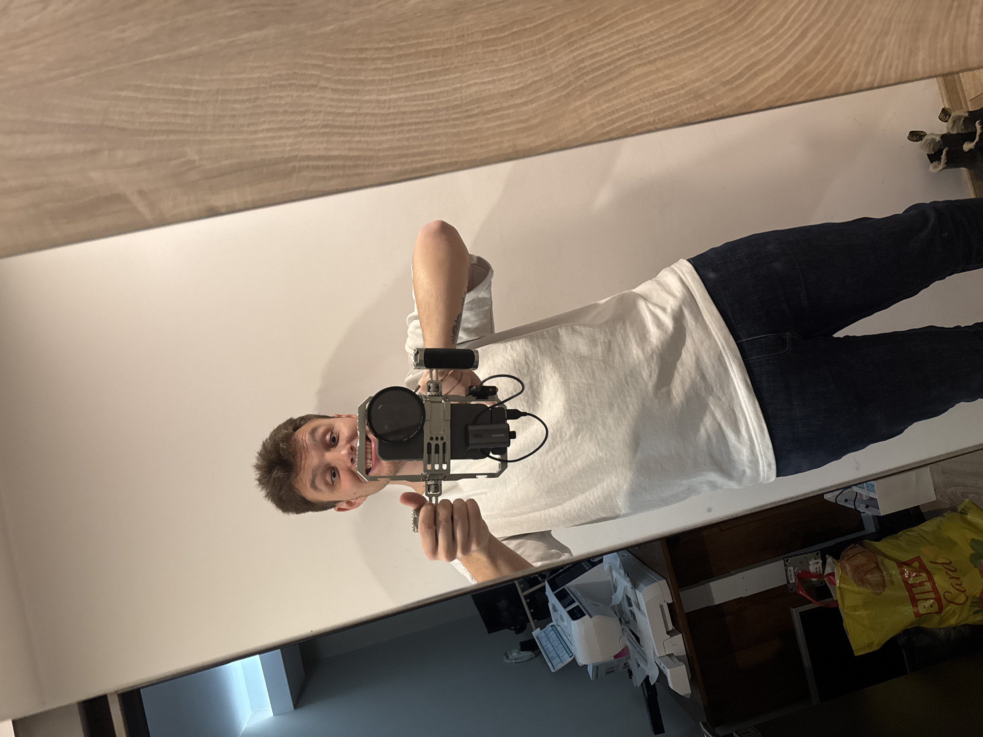 A medium shot depicts a person taking a selfie in a mirror. The person is wearing a white T-shirt and dark jeans. They are holding a camera in a metal frame, equipped with a microphone and external monitor, at eye level. The reflection shows a room with a white wall, a desk or shelf with various items including a printer, papers, and a yellow shopping bag. The background hints at a domestic setting. There is no explicit indication of Plovdiv, Bulgaria.