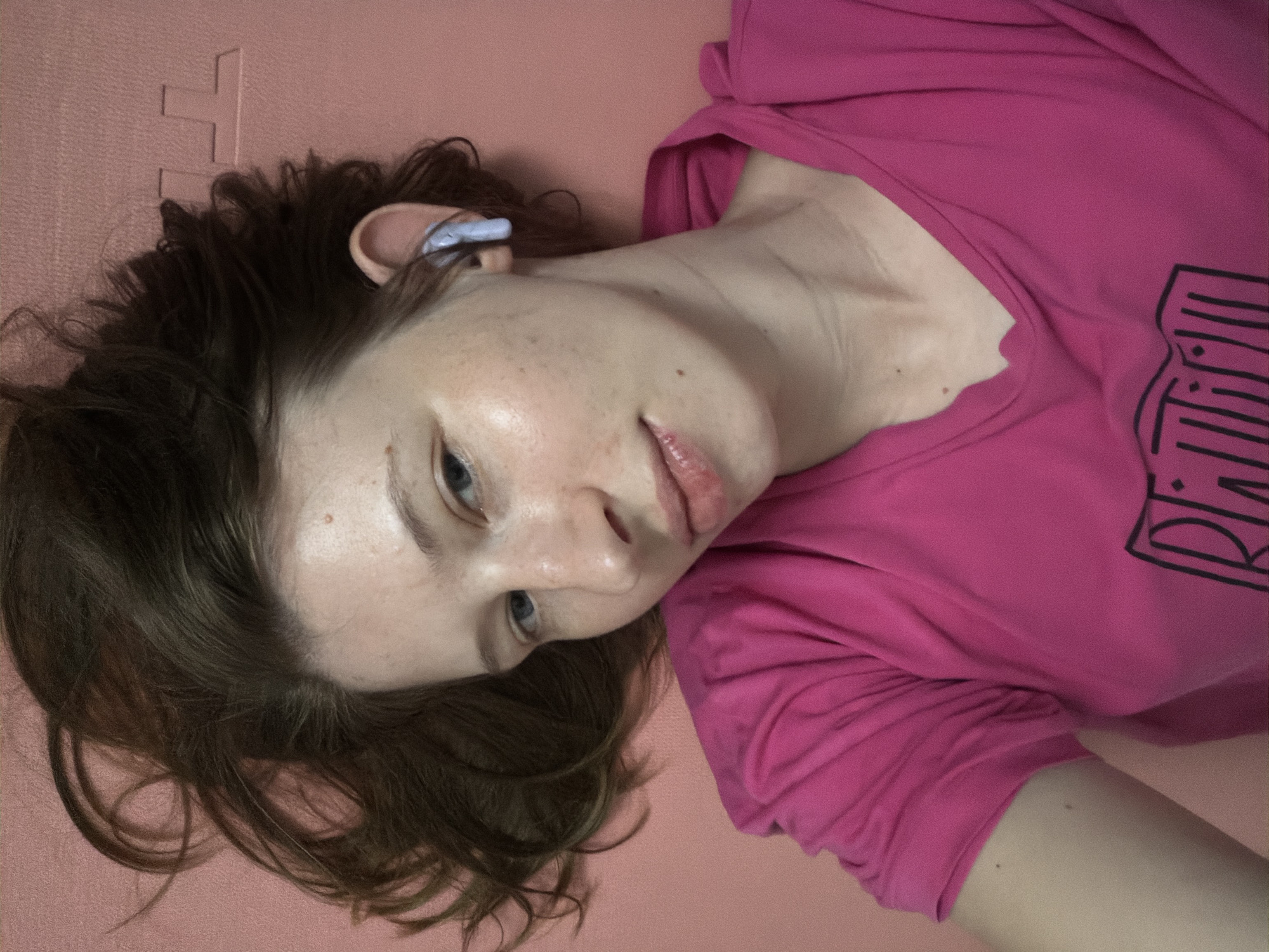 A young woman with light skin, freckles, and light brown hair is lying on her back on a pink mat. She is wearing a bright pink t-shirt and a light blue earbud in her right ear. Her head is tilted to the right, and she is looking upwards towards the camera with a slight smile. The background is a solid pink surface with a subtle textured pattern. The image appears to be a selfie taken from a close-up, overhead perspective. The setting is likely a home or a fitness studio in Dongducheon-si, South Korea.