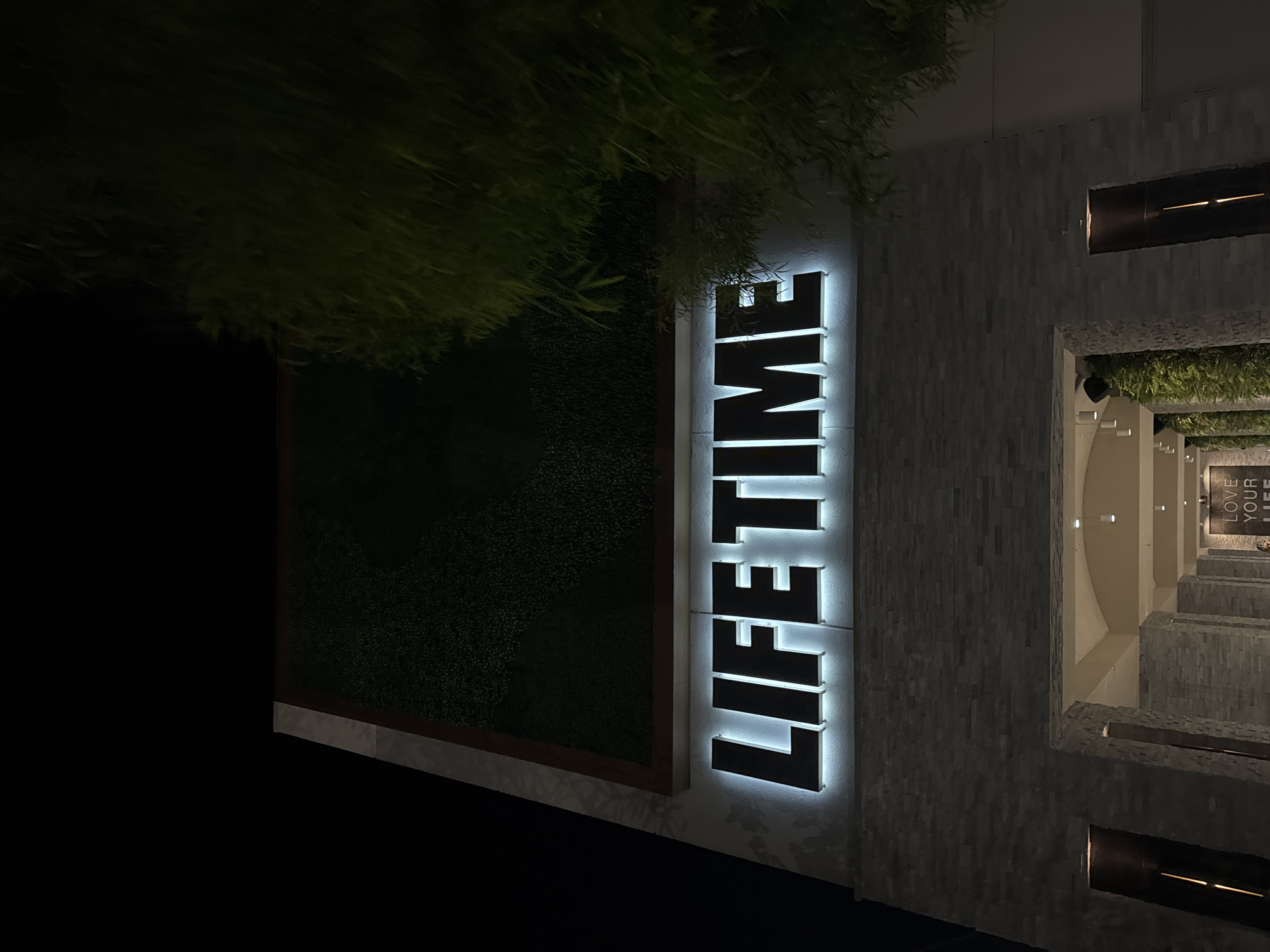The photo shows the exterior of a Life Time fitness center at night. The building is clad in grey stone, and the word "LIFETIME" is displayed vertically in large, illuminated letters. A wall covered in greenery is adjacent to the signage. A glimpse of the entrance reveals interior design features and additional signage. The image was taken in Irvine, California, United States of America.