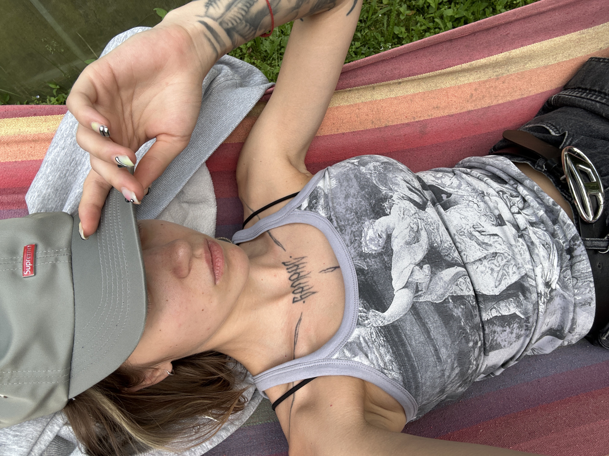 A young woman is lying in a hammock outdoors, wearing a gray baseball cap and a gray tank top with a black and white graphic print. She has a tattoo on her chest and a tattoo on her left arm. She is holding the brim of her cap with her right hand, and her left arm is resting by her side. The hammock is striped in red, orange, yellow, and pink. The background is blurred green grass and a hint of foliage, suggesting an outdoor setting. The image is taken from a high angle, looking down at the woman. The specific location in the city of Kozino, Russia is not identifiable from the image.