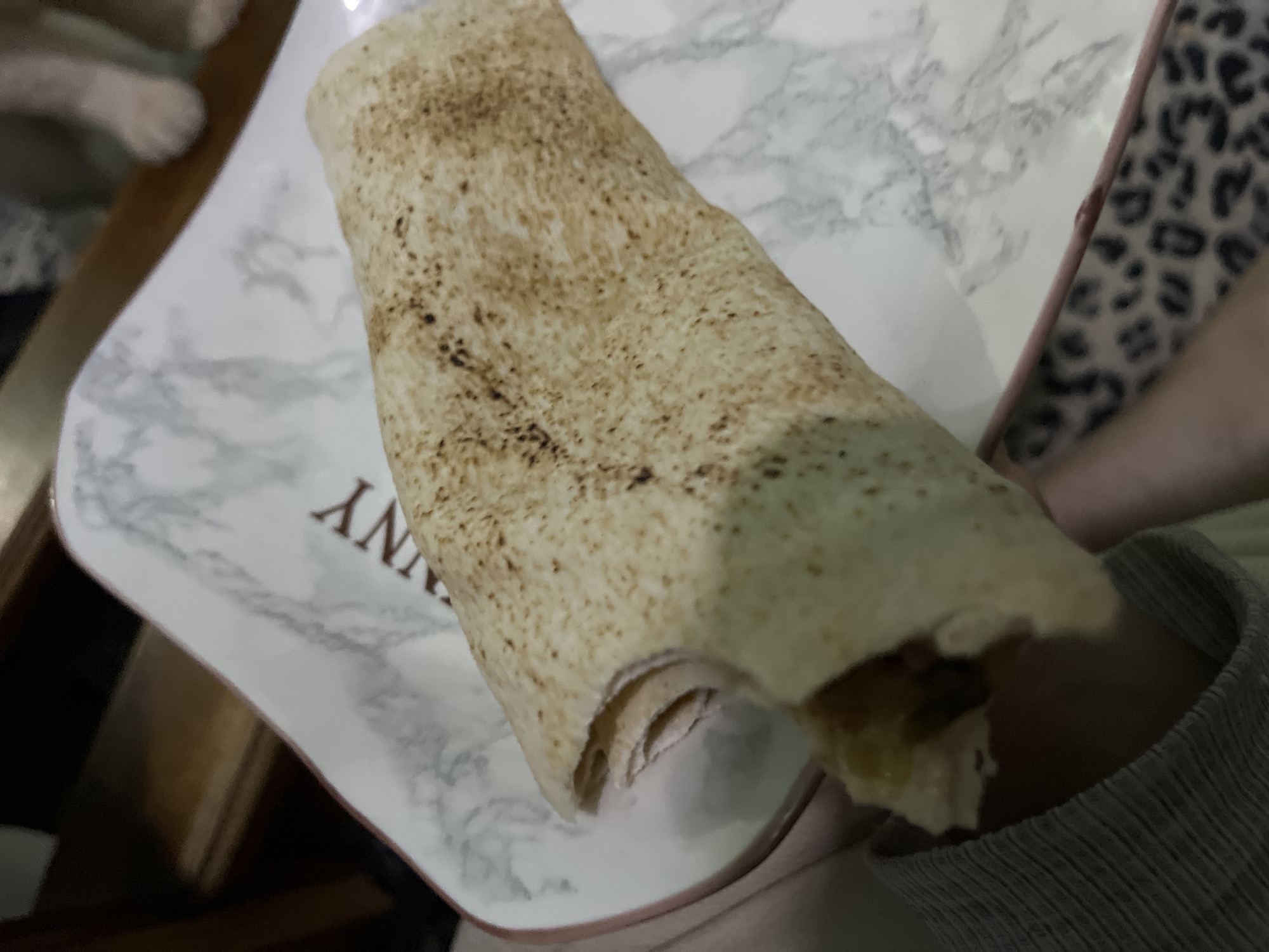 A partially eaten shawarma wrap rests on a white, marbled-patterned plate with dark brown lettering that reads "YANNY". The plate appears to be on a table or held in someone's lap, as indicated by the gray, knitted sweater sleeve visible in the lower right of the frame. The shawarma is browned and slightly charred, with visible filling at the opened end. The background includes a leopard-print pattern on what appears to be a cushion or piece of furniture. Juan Griego, Venezuela, is not discernable from the available visual information.