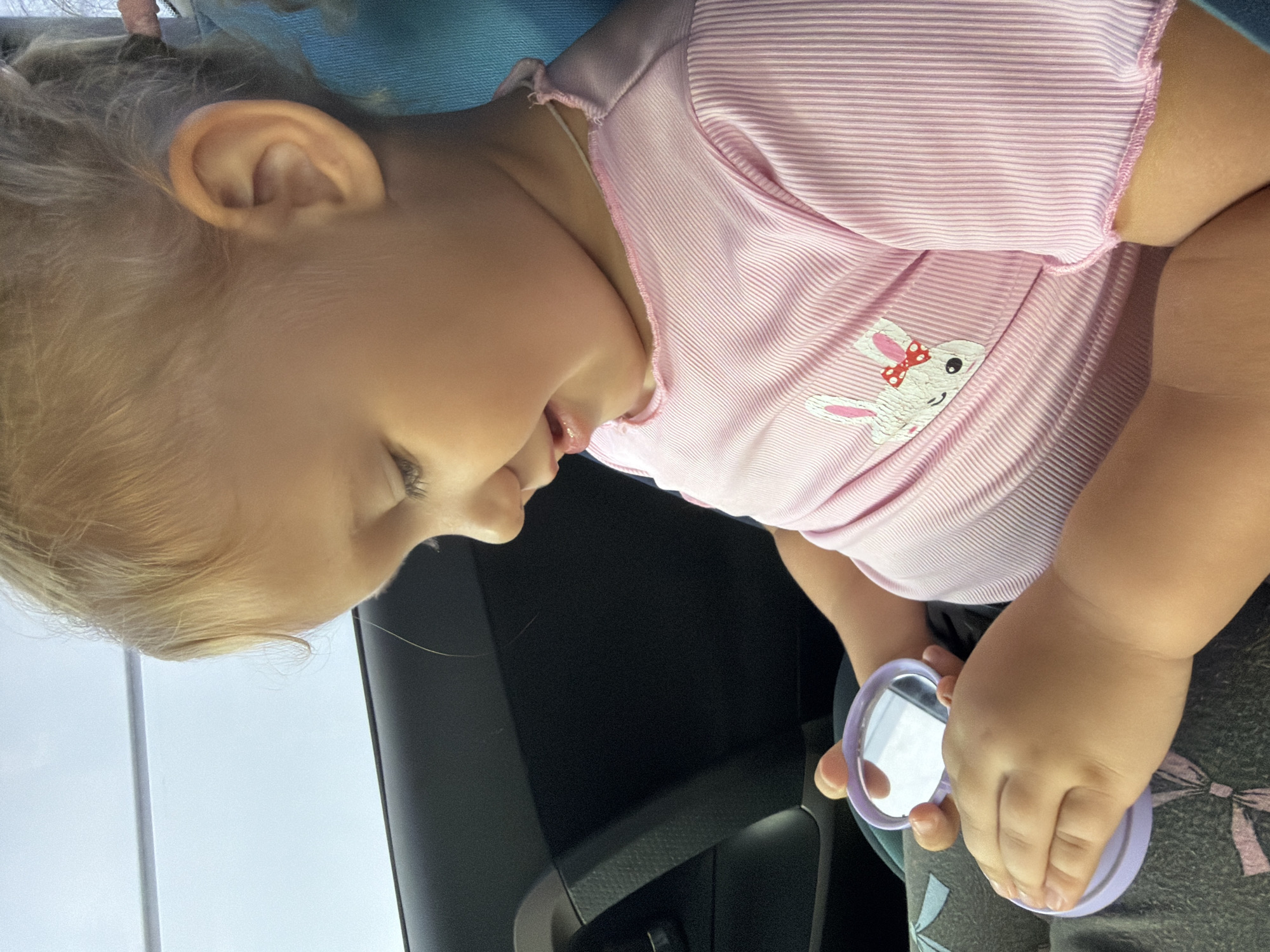 A young child with blonde hair is sitting in a car, looking into a small, purple compact mirror. The child is wearing a pink ribbed t-shirt with a bunny graphic on it. The background shows the interior of a car, including a black door panel and a blue car seat. The image is taken from a close-up perspective, focusing on the child and the mirror. The setting appears to be daylight, possibly during a car ride. The location of Rostov-on-Don, Russia, is not depicted in the image.