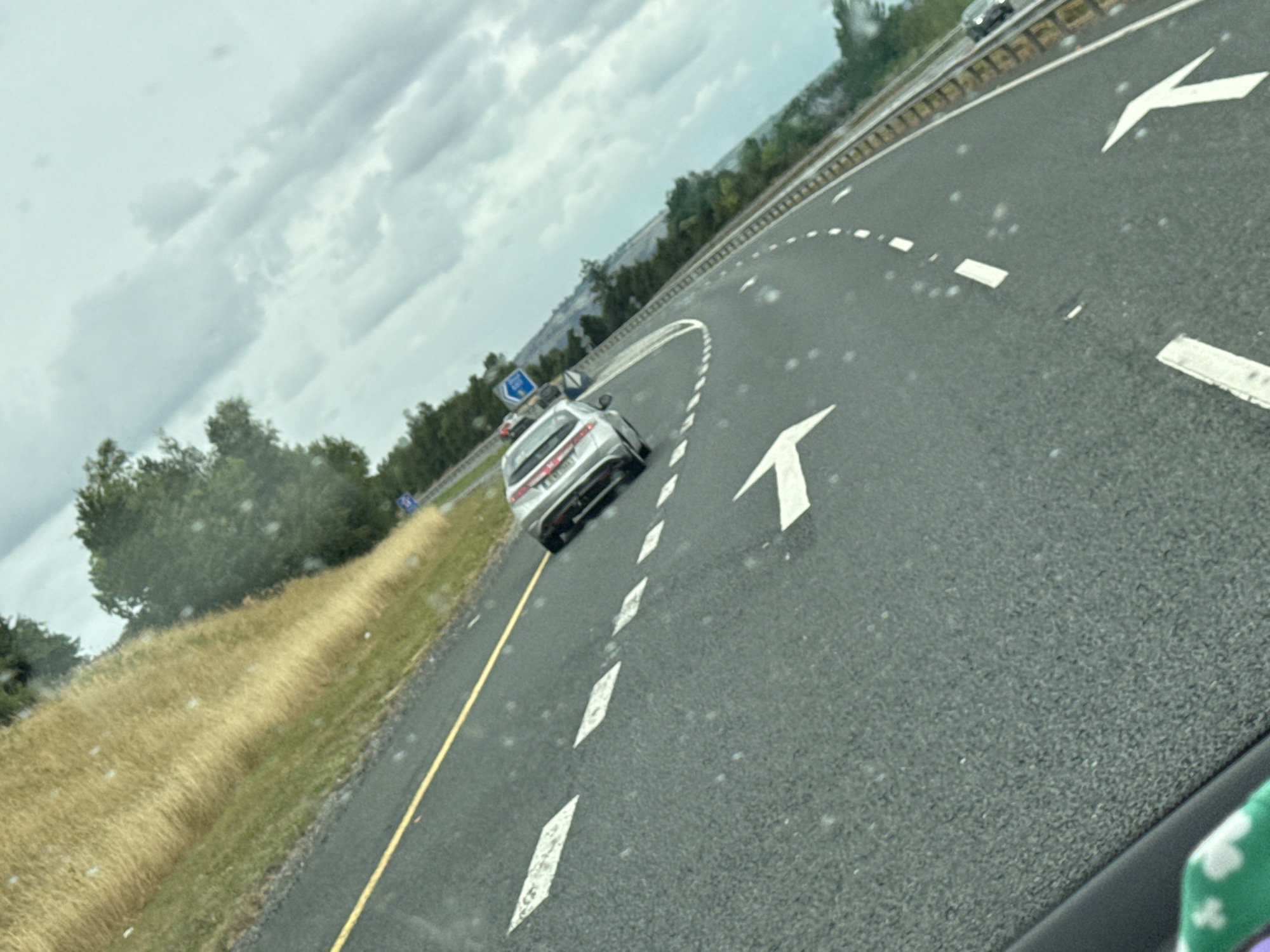 The image depicts a section of a road, likely a highway, with clear lane markings including white arrows and dashed lines. A silver car is visible in the right lane, driving away from the camera. The road curves to the right. On the left side of the road, there is a grassy embankment with dry, tall grass. Trees are present beyond the embankment, and the sky is overcast with clouds. A blue road sign is partially visible in the distance on the left side of the road. The perspective suggests the photo was taken from inside another vehicle. The scene appears to be in a rural or semi-rural setting, consistent with locations like Knockraheen, Ireland, which is known for its scenic roadways and natural landscapes.