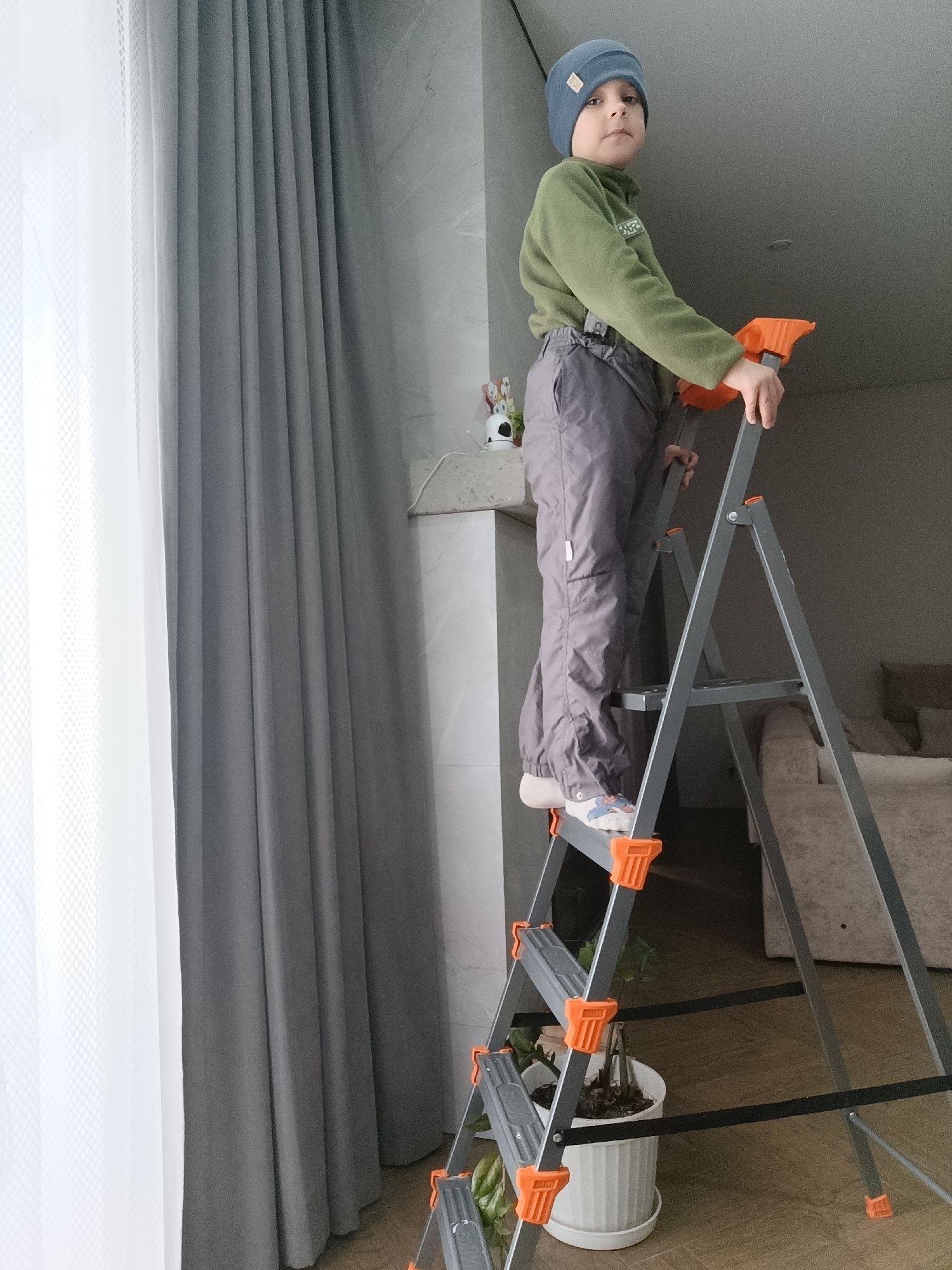 A young child wearing a blue beanie, green sweater, and grey pants stands on a grey and orange aluminum step ladder. The child is gripping the ladder with both hands, looking towards the viewer. The ladder is positioned indoors near a grey curtain and a white fireplace mantelpiece decorated with small figures. A white potted plant with green leaves is visible between the ladder steps. Behind the ladder, a living room area includes a beige sofa. This photo was taken in Chaikovsky, Russia.