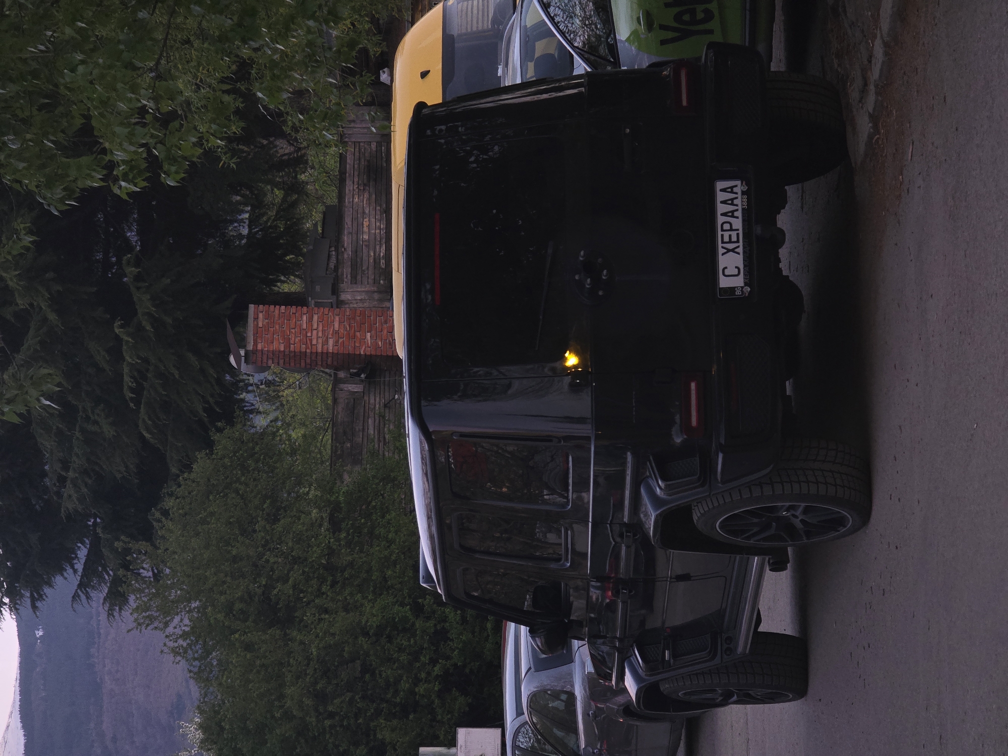 A black Mercedes-Benz G-Wagen is the primary subject, viewed from its rear and slightly to the side. The vehicle is parked on a paved surface, and its spare tire is mounted on the rear door. Its license plate, clearly visible, reads "C XEPAAA 3888" with the "BG" country code for Bulgaria, and 'C' indicating Sofia.
Behind the G-Wagen, a yellow vehicle, possibly a van or small bus, is partially visible, followed by a green vehicle with the partial text "Ye" visible on its side, likely part of a company logo like "Yettel".
The background features dense green foliage from various trees and shrubs, suggesting a natural or park-like area. A brick structure, possibly a chimney or part of a building, and a wooden fence are also visible amidst the greenery. In the far distance, mountains rise under what appears to be a twilight or early evening sky, given the soft, somewhat dim lighting throughout the scene. The overall setting is consistent with a city like Sofia, Bulgaria, situated near mountainous terrain. No explicit human activity is depicted.