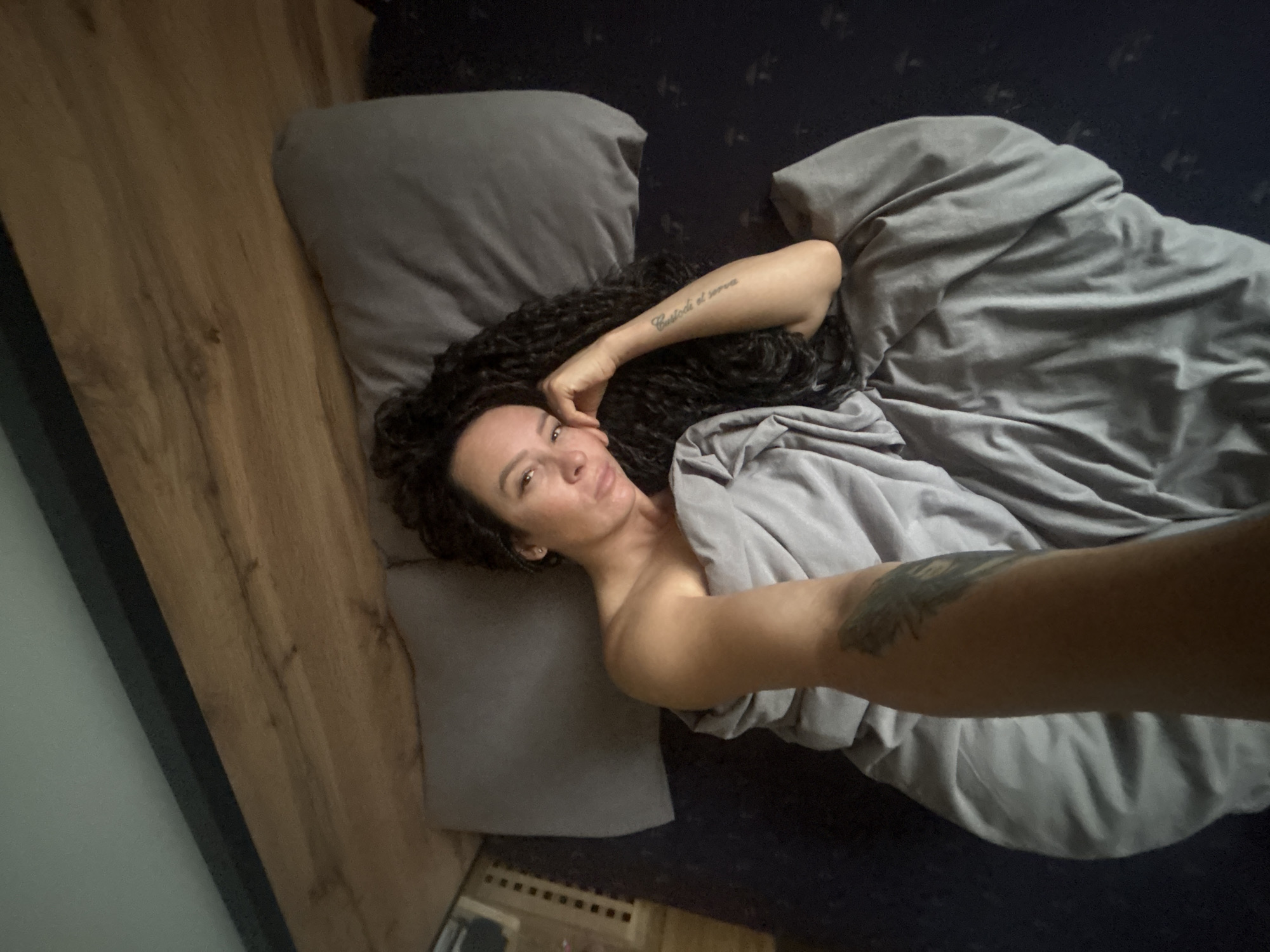 An overhead, slightly tilted photograph, taken in the city of Chaikovsky, Russia, captures a woman lying in a bed. She is positioned on her back, facing the camera with a subtle expression, her head resting on a grey pillow. Her dark, curly hair is spread across the pillow and the dark-colored fitted sheet. A grey duvet covers her body up to her shoulders, exposing her right arm, which is bent with her hand near her face. A script tattoo is visible on her right forearm, and a darker, feather-like tattoo is partially visible on her left forearm beneath the duvet. The bed features a light wooden planked headboard along the right side of the frame. The bed's fitted sheet is dark blue or black with a faint, repeating pattern. The background includes a light greenish-grey wall in the upper right, bordered by a thin dark vertical line, and a dark blue surface with a small, light-colored slatted object in the upper left. The setting is an indoor bedroom.