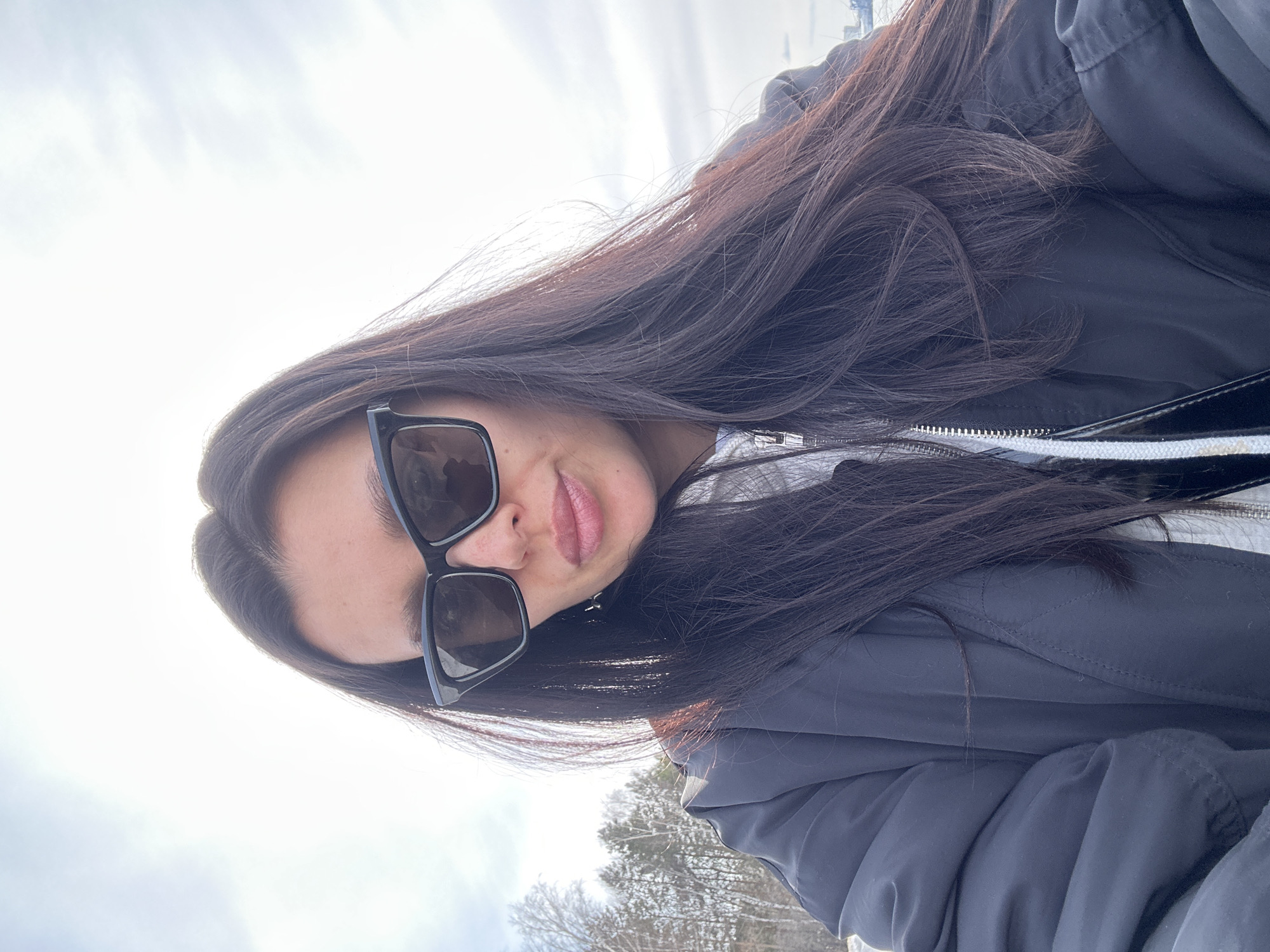 A selfie of a woman wearing sunglasses and a dark jacket. Her long, dark hair is spread around her face and shoulder. The background shows a bright sky with what appears to be a winter landscape with bare trees. The picture was taken near the city of Chaikovsky, Russia.