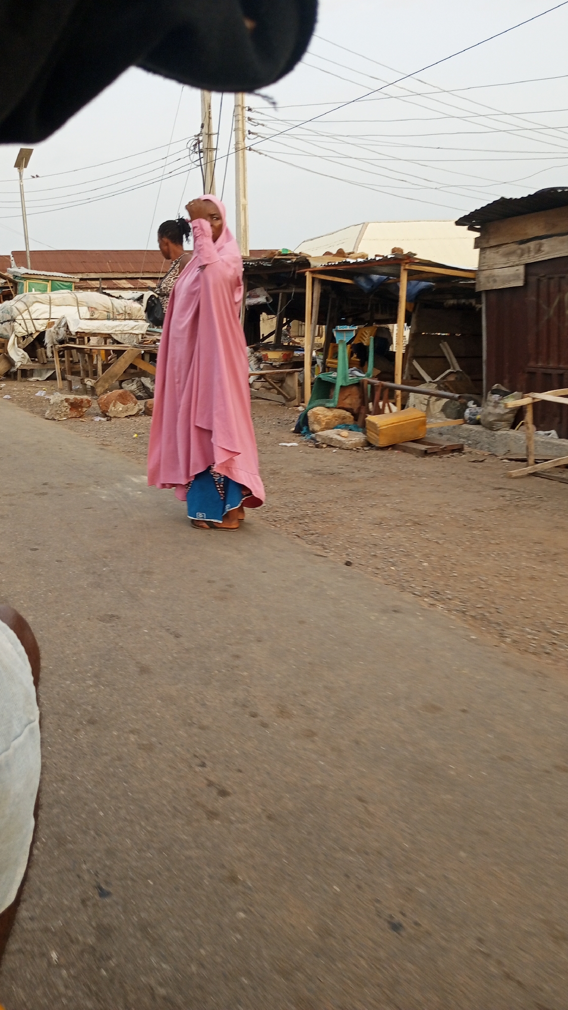 The image shows two individuals on a street in an urban environment. The foreground is dominated by a road surface that appears to be paved but somewhat worn. The main subject of the image is a woman wearing a long, flowing pink outer garment or robe, with a blue lower garment visible underneath. She is adjusting her head covering with her right hand. Beside her, partially obscured, is another person with a dark dress and her hair in braids. In the background, there are several makeshift structures or market stalls, some covered with tarps or canvas, others open. Debris, rocks, and other miscellaneous items are scattered around the stalls. Overhead, electrical wires span across the sky, connected to utility poles. The location appears to be a developing city with informal market structures alongside a paved road. The city is Unknown, Unknown.