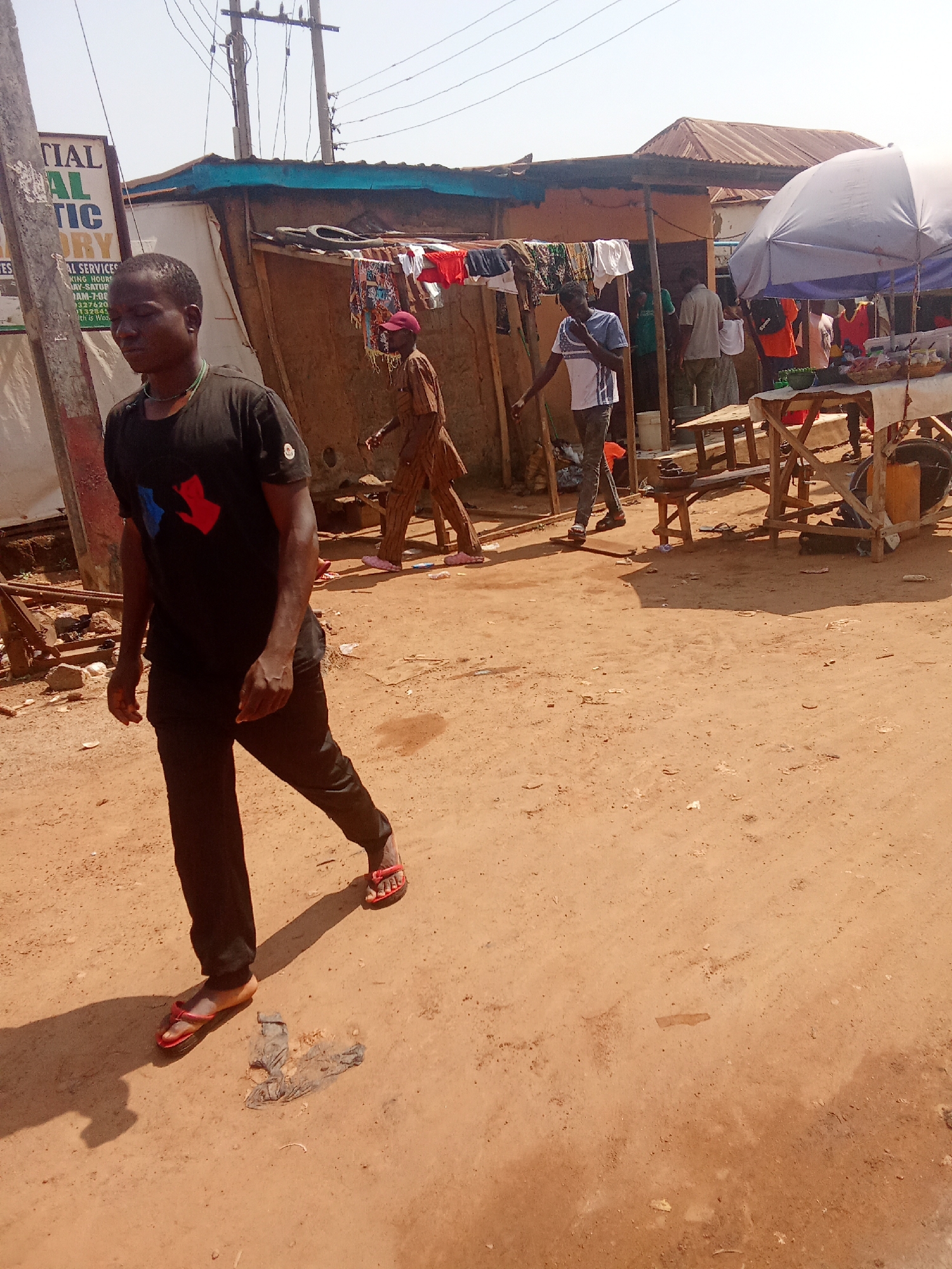 In Kagini, Nigeria, a street scene unfolds under a bright sky. A man in a black t-shirt and black trousers walks from left to right, his gait suggesting purpose. His t-shirt bears a red and blue emblem on the chest. He wears red flip-flops. Behind him, another man, possibly a vendor, stands near a stall. He is dressed in a brown traditional outfit and a red cap, and appears to be attending to goods displayed on a wooden table under a large umbrella. In the background, a cluster of makeshift structures with corrugated iron roofs and fabric coverings line the street. Laundry hangs on a line strung between two structures. Other individuals are visible in the distance, suggesting a bustling marketplace or community area. The ground is a dusty, reddish-brown earth, littered with debris. The overall impression is one of everyday life in a semi-urban or rural Nigerian setting.