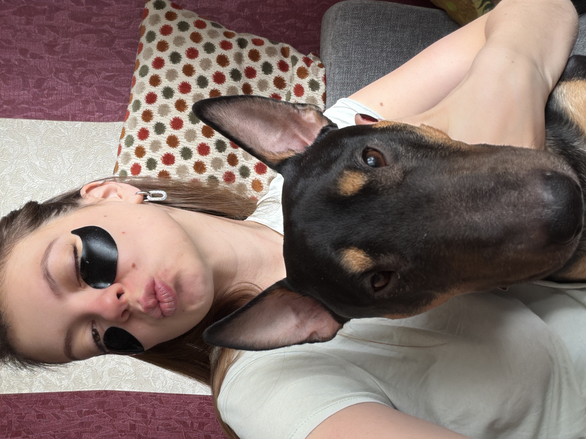 This is a close-up, high-angle indoor photograph capturing a relaxed moment between a young woman and a dog in a home setting in Khimki, Russia. The image shows the subjects positioned horizontally on what appears to be a bed or sofa.
The primary subjects are a young woman with long brown hair and a dark-colored dog. The woman is lying down, wearing a light-colored t-shirt. She has black, crescent-shaped under-eye patches on her face, suggesting a self-care routine. Her lips are pursed in a playful pout, and her eyes are partially closed, contributing to a whimsical or affectionate expression. A silver-toned ear cuff embellished with small rhinestones is visible on her left ear.
The dog's head is prominently featured close to the woman's face, indicating a close bond. It has short black fur with tan markings around its muzzle and eyes, and one of its large, triangular ears is clearly visible. The dog's brown eyes are directed towards the woman or the camera with a gentle and curious gaze.
The background elements include a textured off-white surface beneath the woman, resting on a textured reddish-purple base, likely blankets or sheets. A patterned pillow with red, green, brown, and off-white polka dots is behind her head. To the right, a gray sofa armrest or cushion is visible, contributing to the domestic and cozy atmosphere. The scene suggests a quiet moment of companionship and relaxation. No text is visible in the image.