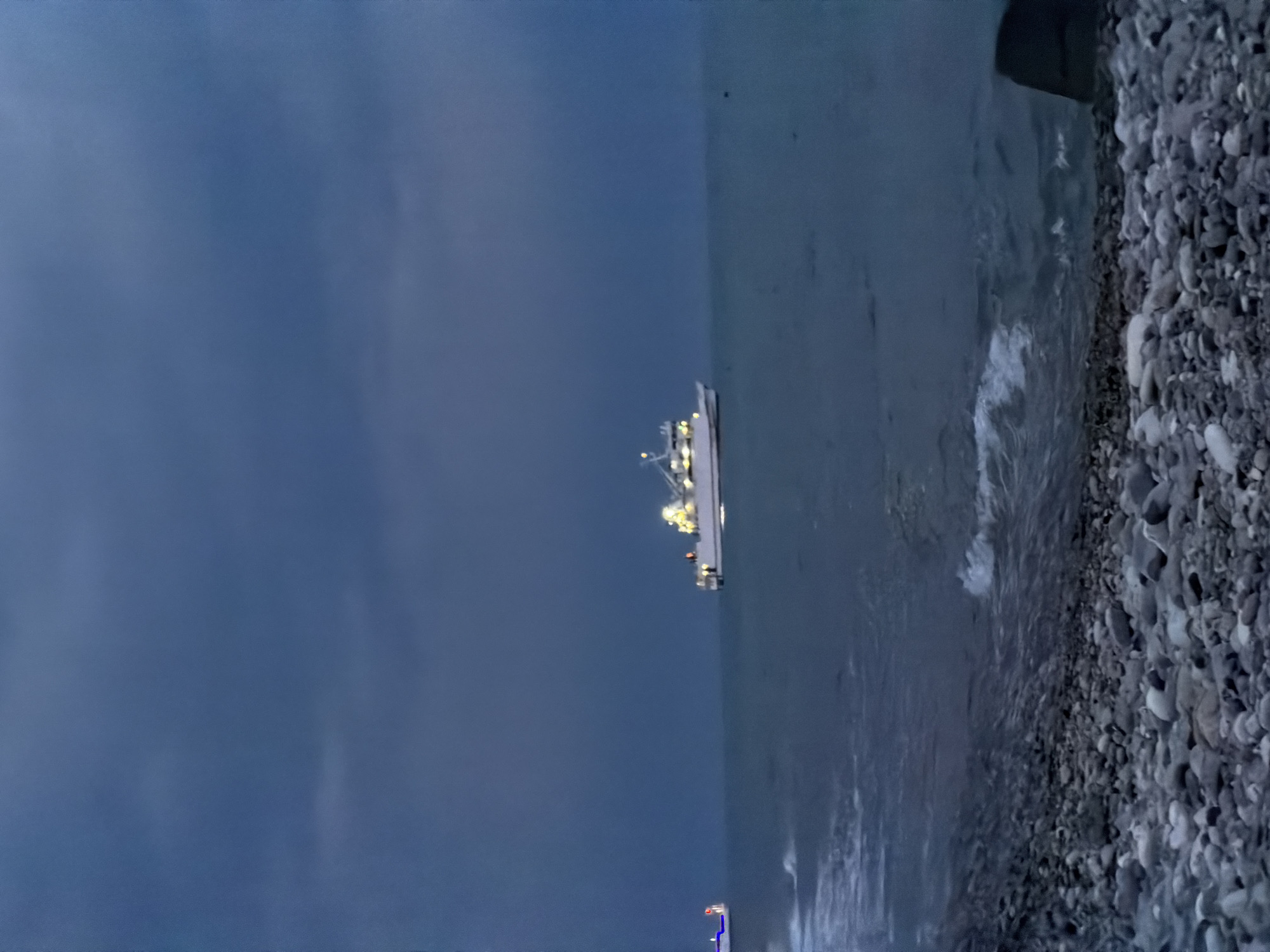 A wide shot captures a serene evening scene on a rocky beach in Sochi, Russia. The sky is a deep, moody blue, hinting at twilight or approaching dusk. Two ships are visible on the calm sea. The larger ship, positioned in the mid-right of the frame, is white with lit windows and appears to be a passenger or research vessel. The smaller ship on the left is also lit, with blue lights visible. The foreground of the image is dominated by a pebble-strewn beach, with gentle waves lapping at the shore. The overall atmosphere is tranquil and atmospheric.