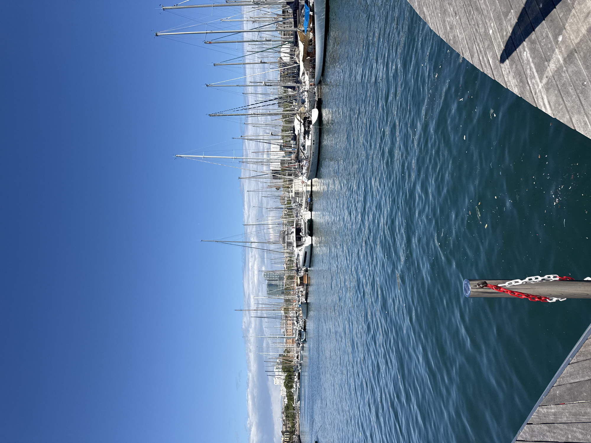 The image depicts a harbor scene in Barcelona, Spain, likely Port Vell, with numerous sailboats and yachts moored along a wooden dock. The sky is clear and blue. The masts of the sailboats reach high into the sky. A segment of the wooden dock is visible in the foreground, including a wooden post with a red and white chain attached. The water is calm and reflects the sky and boats. A line of buildings is visible along the horizon.