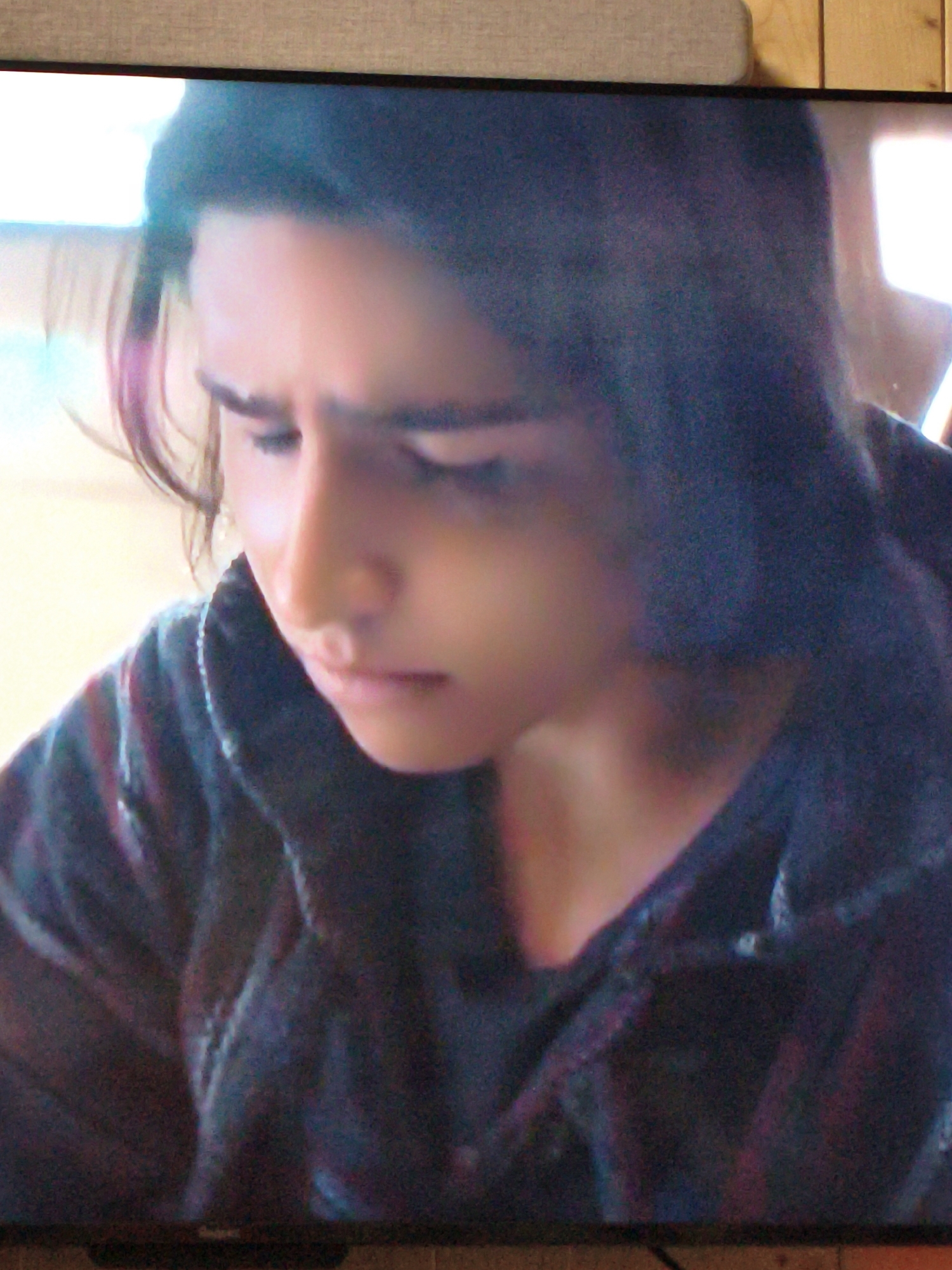 The image is a close-up, grainy, and somewhat blurry photograph of a person with long, dark hair looking down. The person is wearing a dark hooded jacket. The lighting suggests the scene is indoors, possibly in front of a window or light source that is out of frame to the left, casting a bright light on the left side of their face. The background consists of a textured surface and what appears to be wooden paneling. There is no discernible information about the location being Vladivostok, Russia. The image is an indoor portrait, likely taken with a camera or phone, and appears to be a still from a video due to the display frame at the bottom.