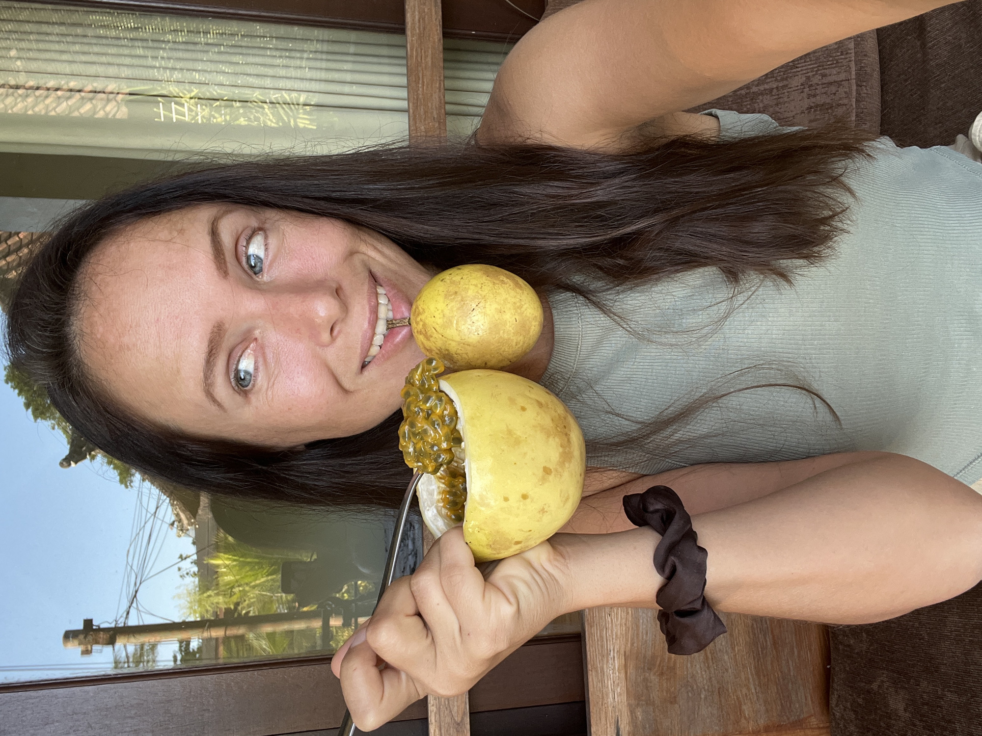 A young woman with long, dark hair and blue eyes smiles brightly, looking directly at the camera. She holds a halved yellow passion fruit with a spoon, poised to eat its orange, seed-filled pulp. Another whole yellow passion fruit rests near her shoulder. She wears a light green, ribbed top, and a dark brown scrunchie is on her right wrist. Her expression conveys joy and pleasantness.
The setting appears to be indoors, but she is positioned near a large window or glass door. The background, visible through and reflected in the glass, depicts an outdoor tropical environment. Elements such as lush green foliage, trees, wooden architectural details, and utility poles with overhead wires are discernible under a bright, clear sky. This suggests a sunny daytime, likely morning or early afternoon. No visible text is present in the image. The presence of tropical fruits and the outdoor environment are consistent with the provided location context of Ubud District, Indonesia.