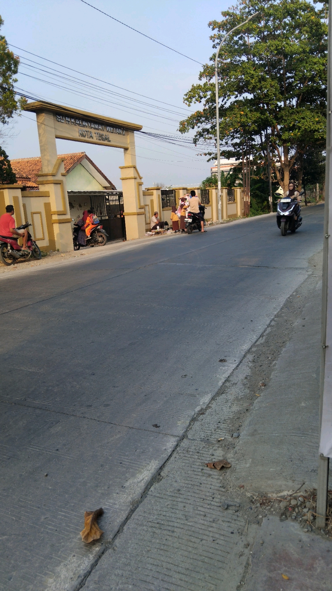 The image captures an outdoor scene on a street in Tegal, Indonesia, likely in front of a school or community building, as indicated by the large archway with the inscription "SUNAN KALIJAGA TEGAL" and "KOTA TEGAL". The sky is clear and light blue, suggesting daytime.
In the foreground, the road surface is grey asphalt with distinct textured lines. A single dry brown leaf is visible on the road. A concrete curb separates the road from a dirt and gravel embankment.
The midground features several people and motorcycles near the entrance of a building. On the left, a person in a red shirt sits on a red motorcycle. Further to the right, a group of individuals are gathered. Some are sitting on the ground, while others are standing or on motorcycles. The building behind them has a tiled roof and a yellow and brown facade with a black metal gate.
In the background, a tall tree with green foliage stands to the right of the archway. A street lamp is also present near the tree. A person on a dark-colored motorcycle is riding on the road, moving away from the viewer. The overall impression is a snapshot of daily life on a street in Tegal.